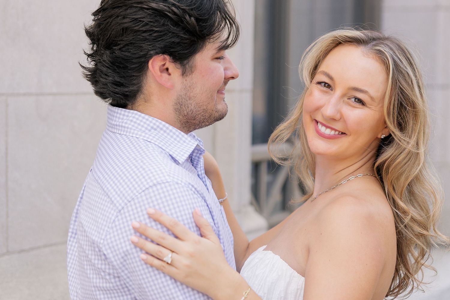 Smiling blonde woman in strapless dress embraces dark-haired man in blue shirt during romantic engagement photo session.