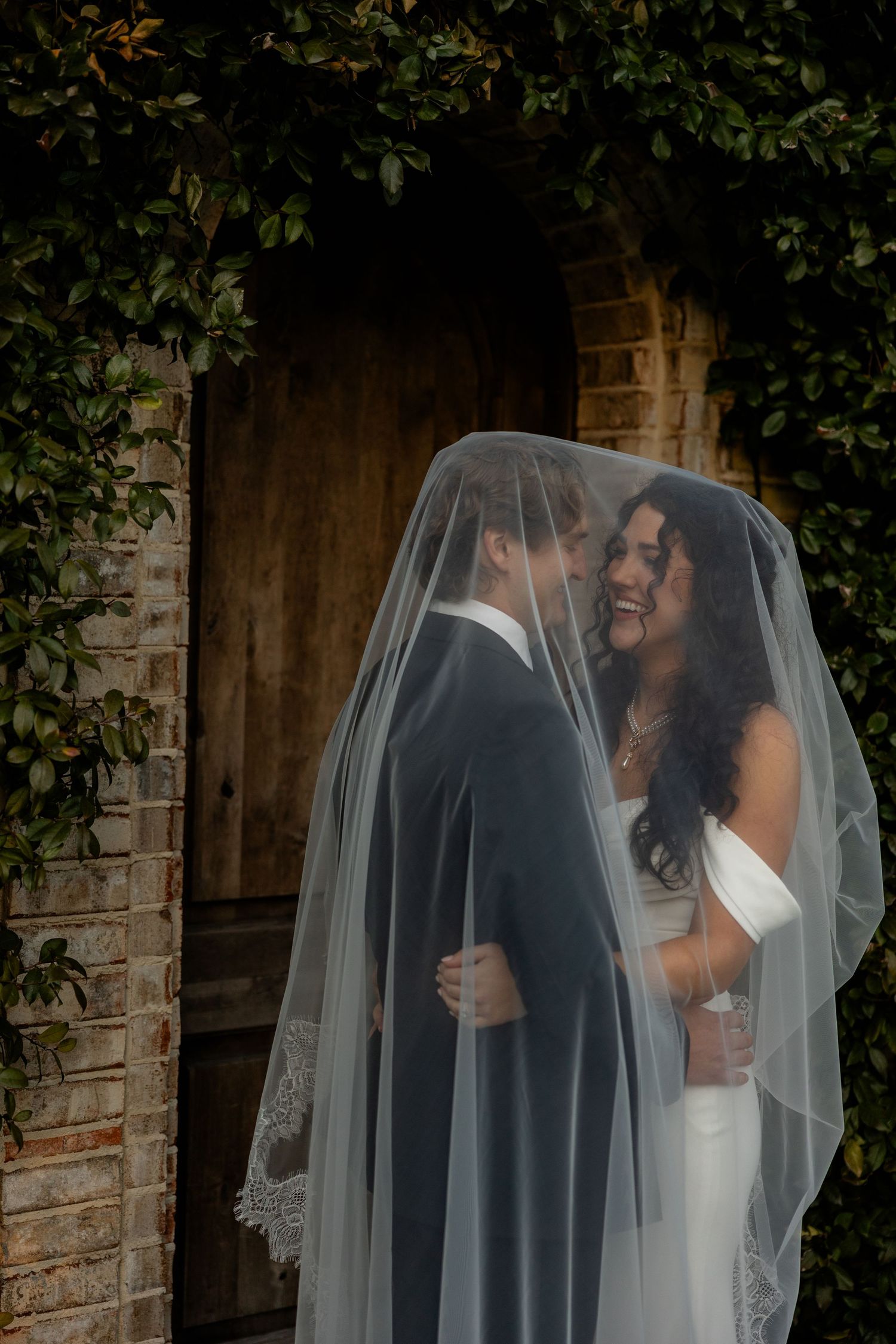 A couple shares an intimate moment under a wedding veil against a rustic brick backdrop with greenery.