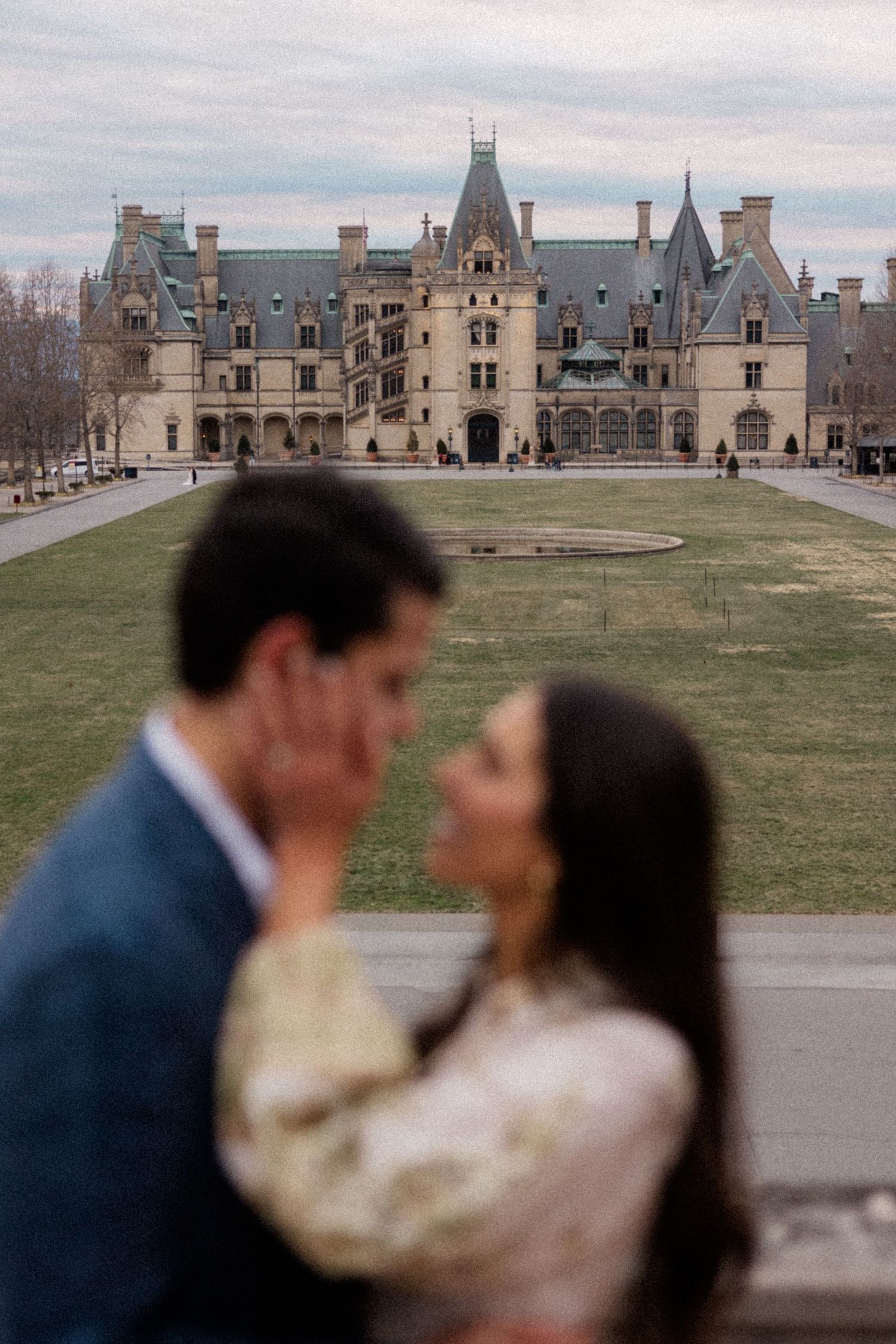A couple stands in front of the majestic Biltmore Estate mansion with its French Renaissance architecture on a cloudy day.