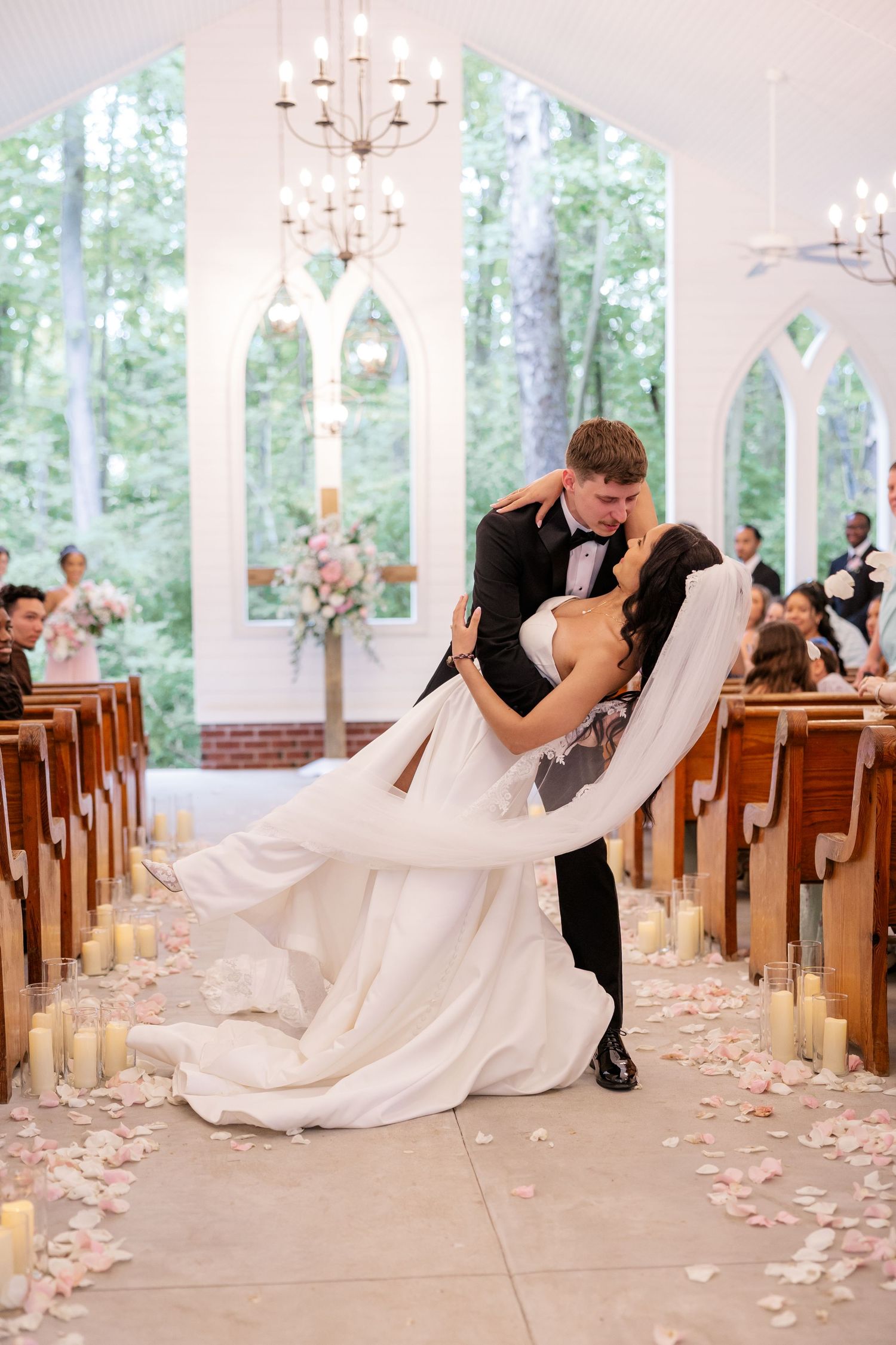 Romantic wedding couple dips and kisses in a white chapel with wooden pews and rose petals.