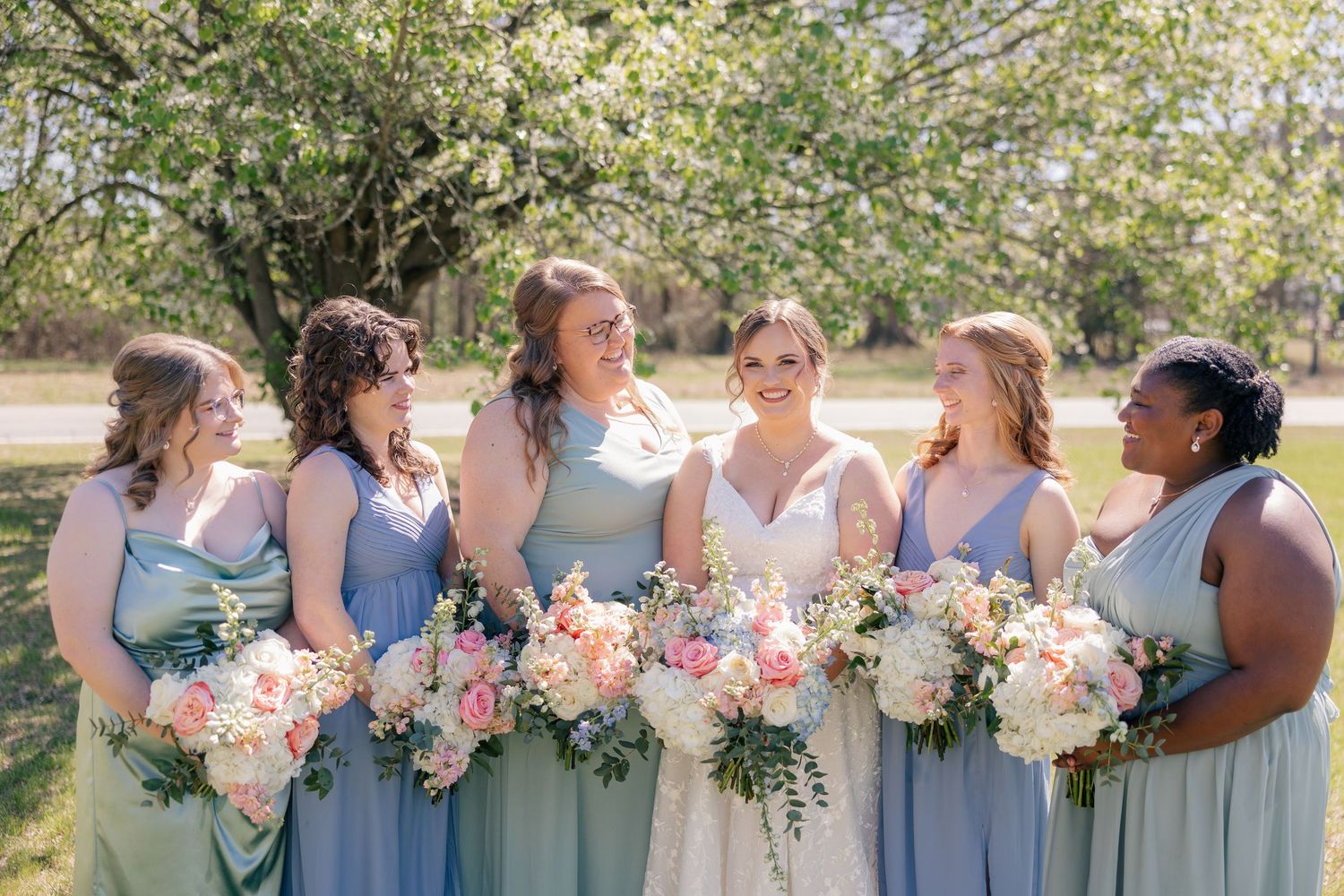 A group celebrates outdoors holding elegant bouquets of pink and white flowers in matching blue dresses.