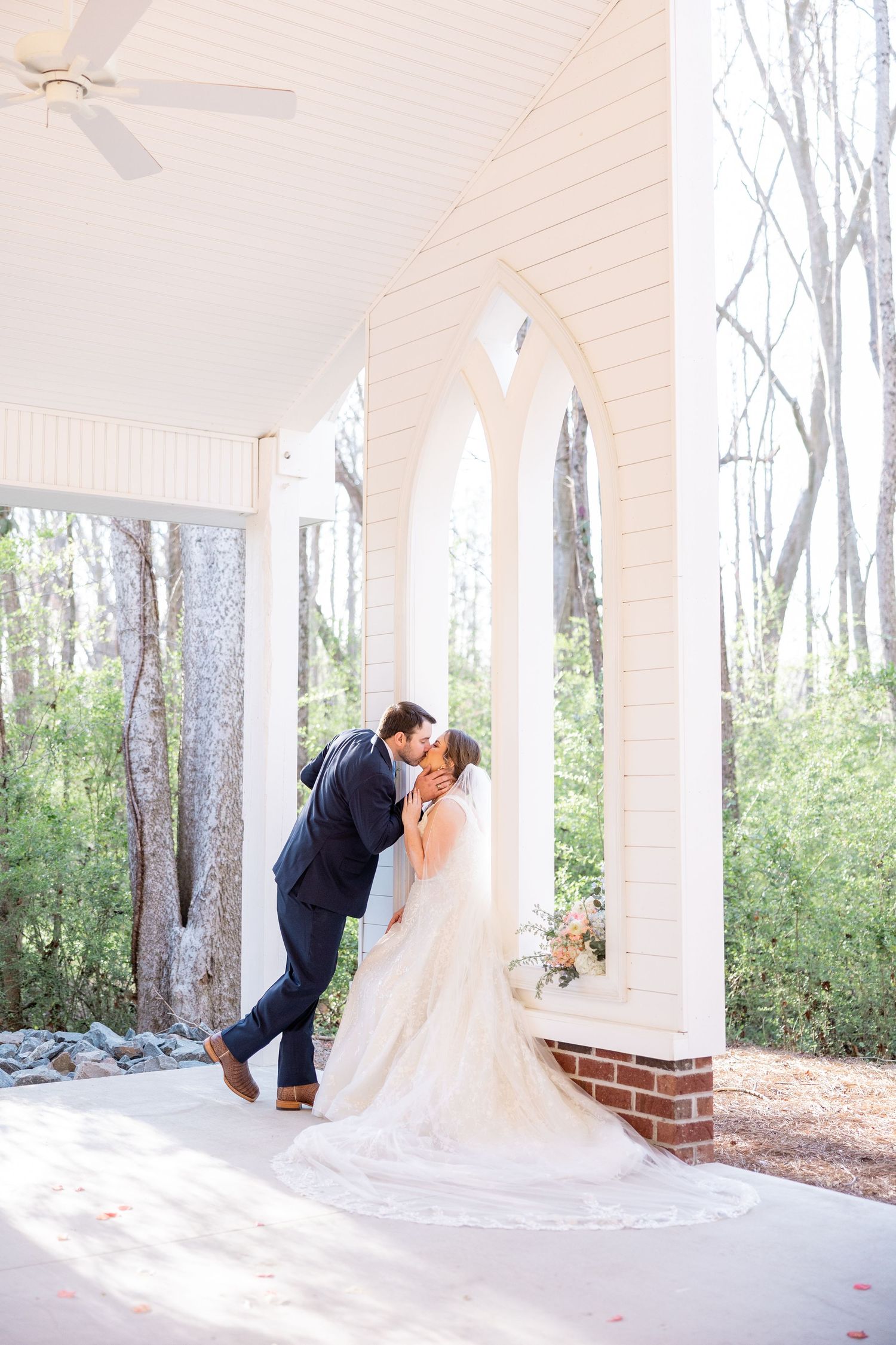Wedding couple shares romantic moment on white columned porch surrounded by trees.