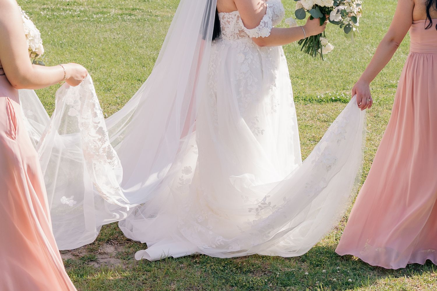 Wedding dress train and blush pink bridesmaid dresses captured during an outdoor ceremony.