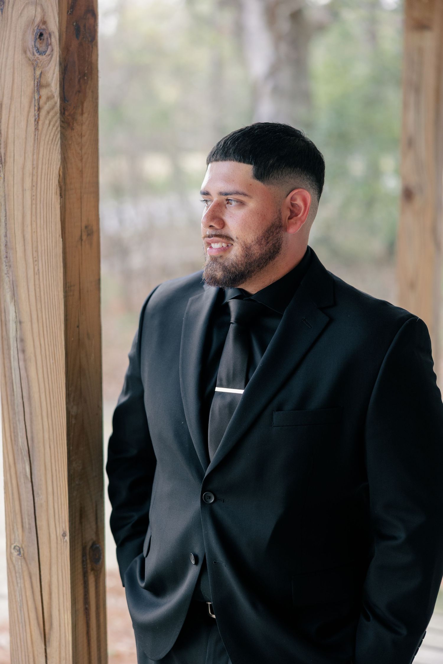 A formal portrait of someone in a black suit and tie standing against a rustic wooden backdrop outdoors.