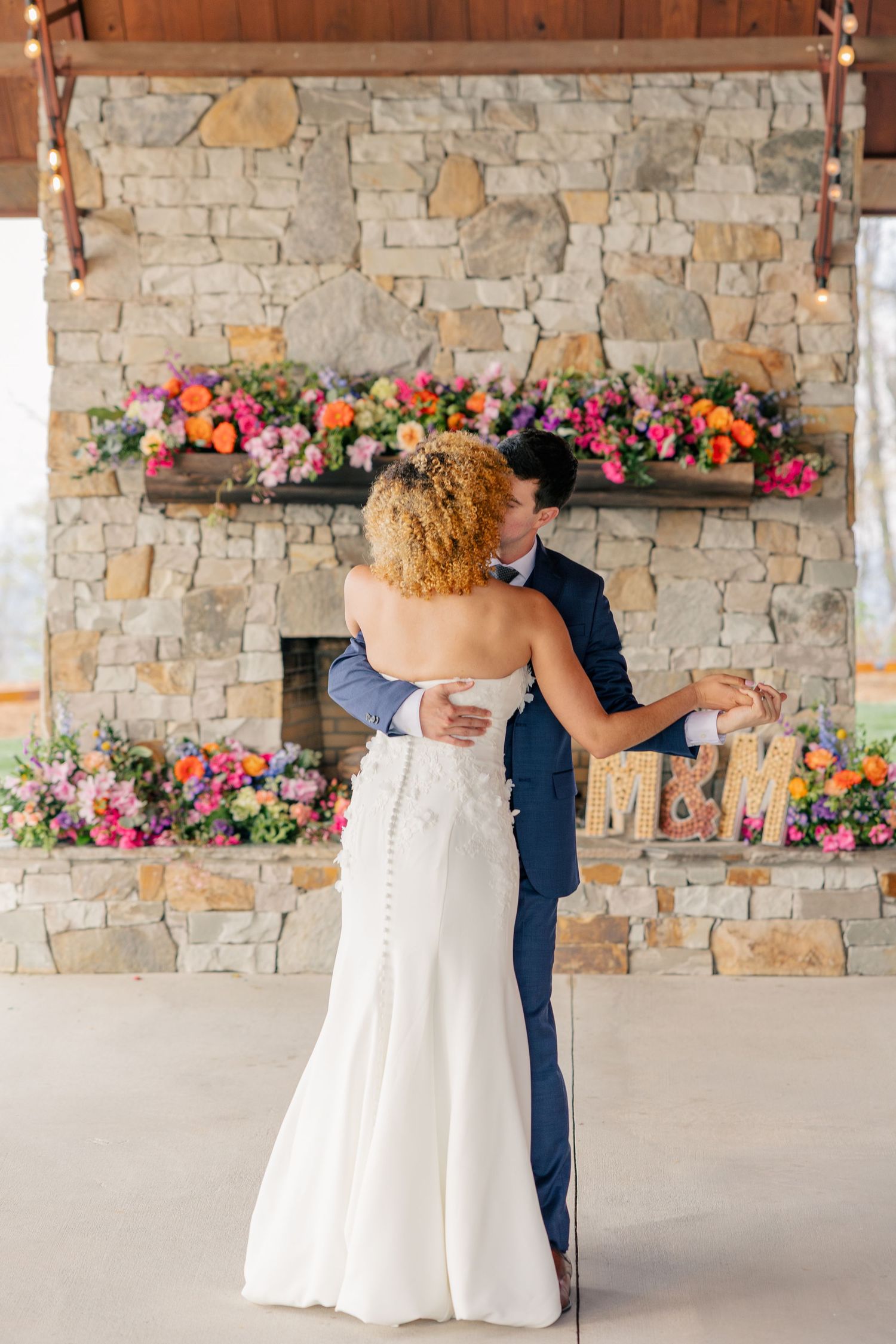 Bride and groom share first dance in front of a stone fireplace adorned with vibrant colorful flowers.