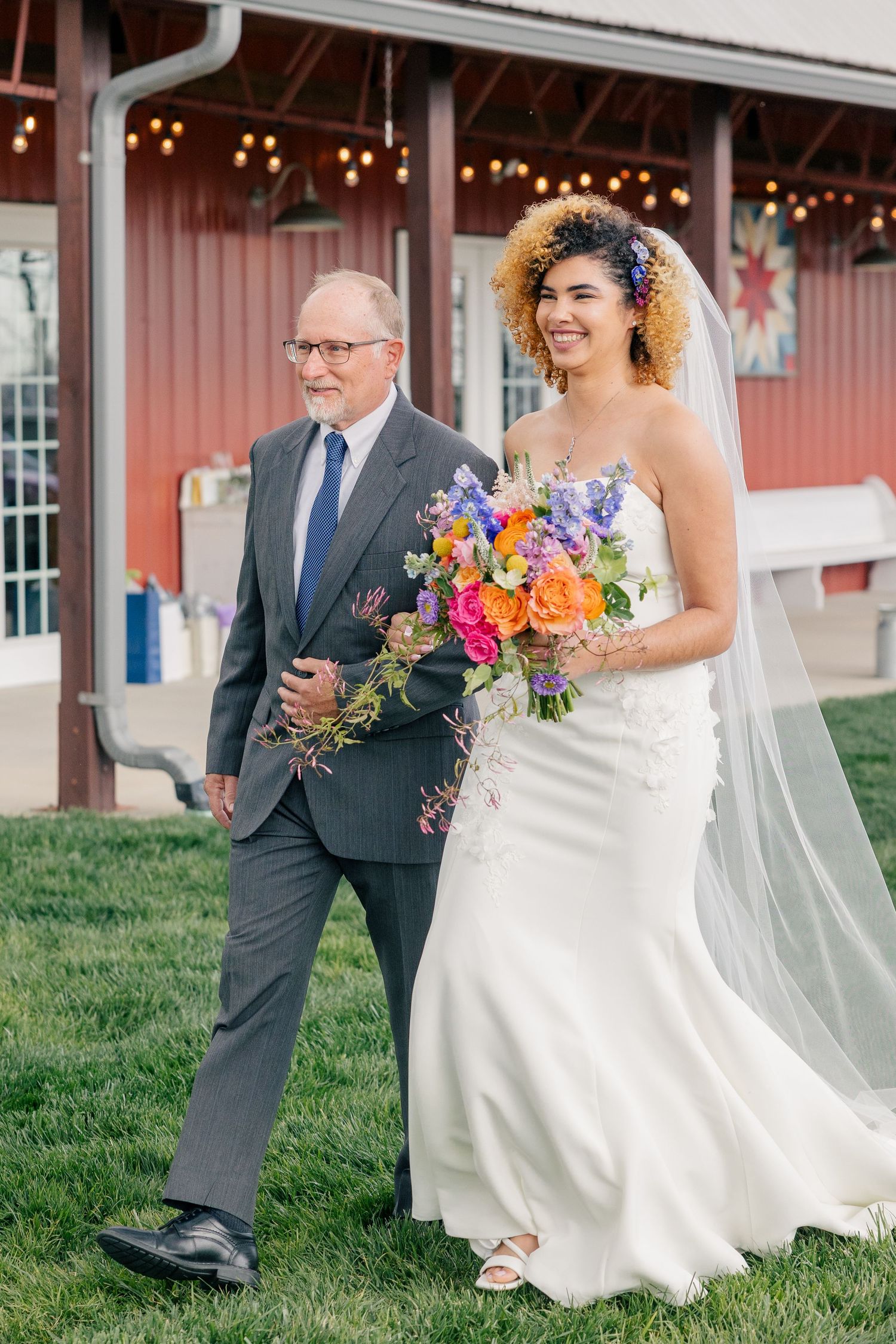 Smiling bride in strapless white gown carrying vibrant wildflower bouquet while walking with her father.