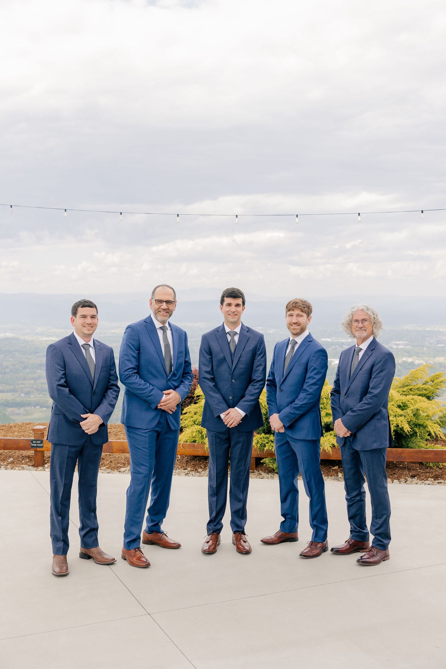 Groom and four groomsmen in navy blue suits pose on outdoor terrace with panoramic sky backdrop.