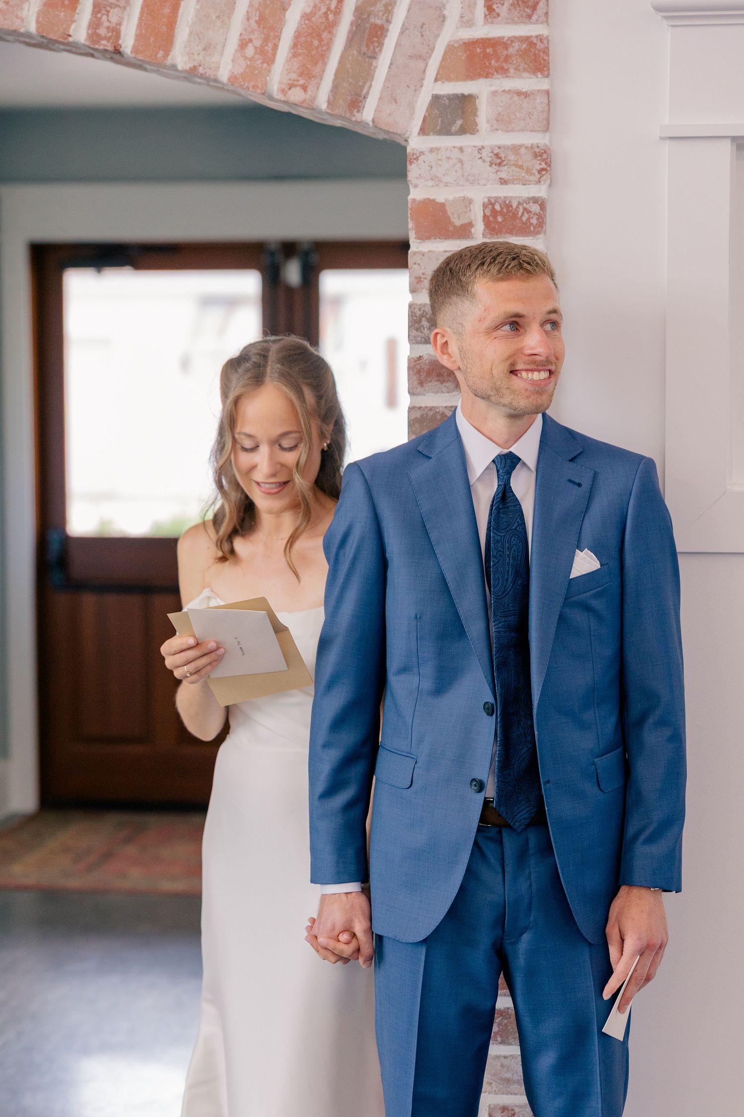 Bride reads vow letter while holding groom's hand before wedding ceremony in blue suit.