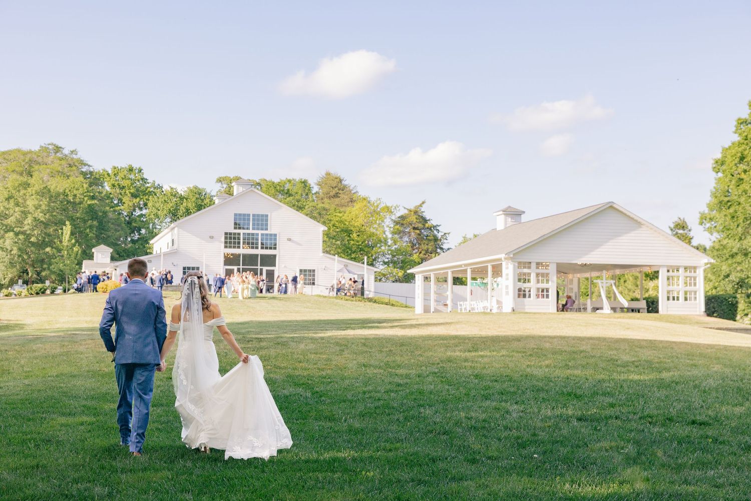 Newlyweds walk hand in hand across green lawn toward white barn wedding venue in warm afternoon light.