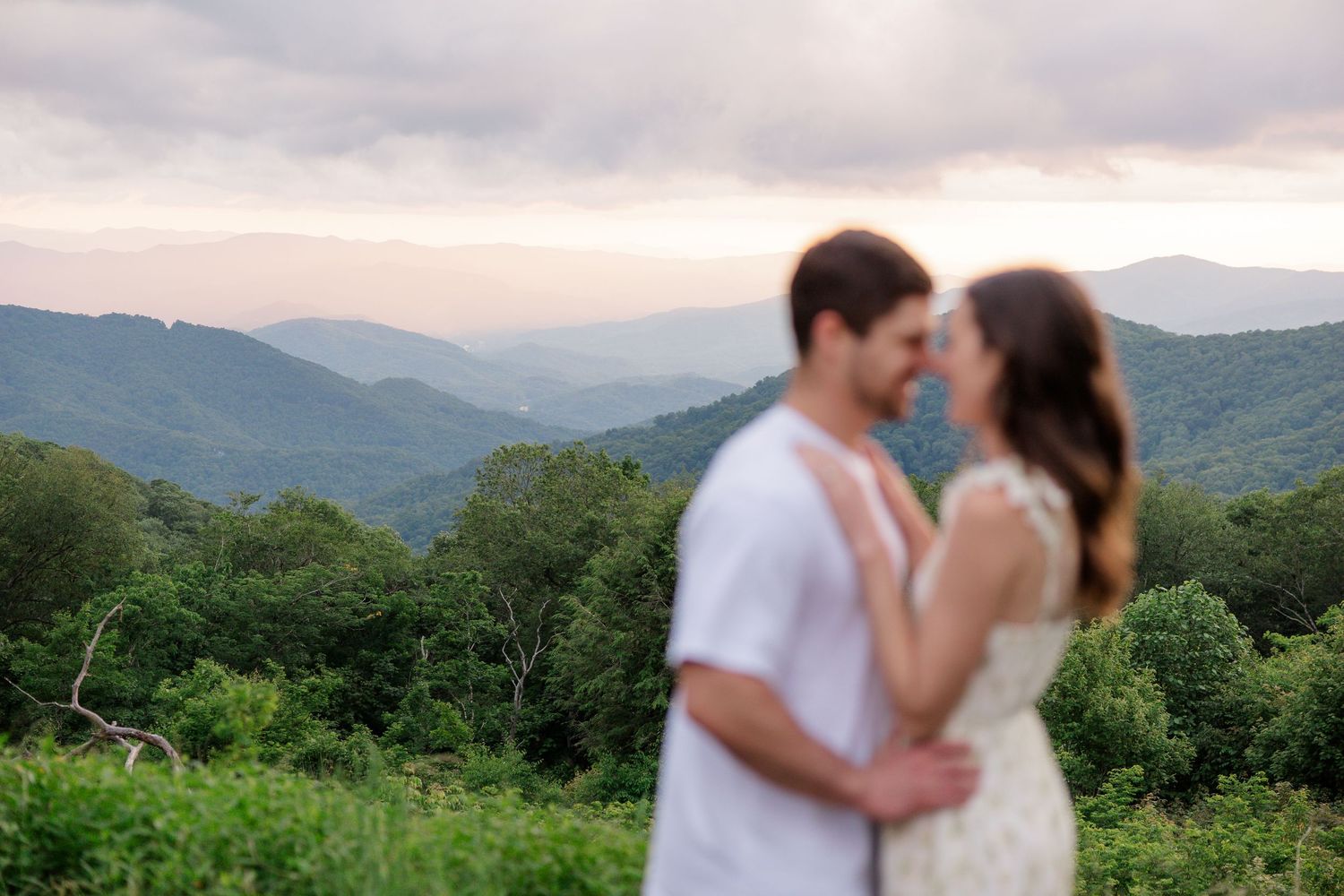 A romantic couple embraces while standing in a mountain meadow with misty peaks in the background.