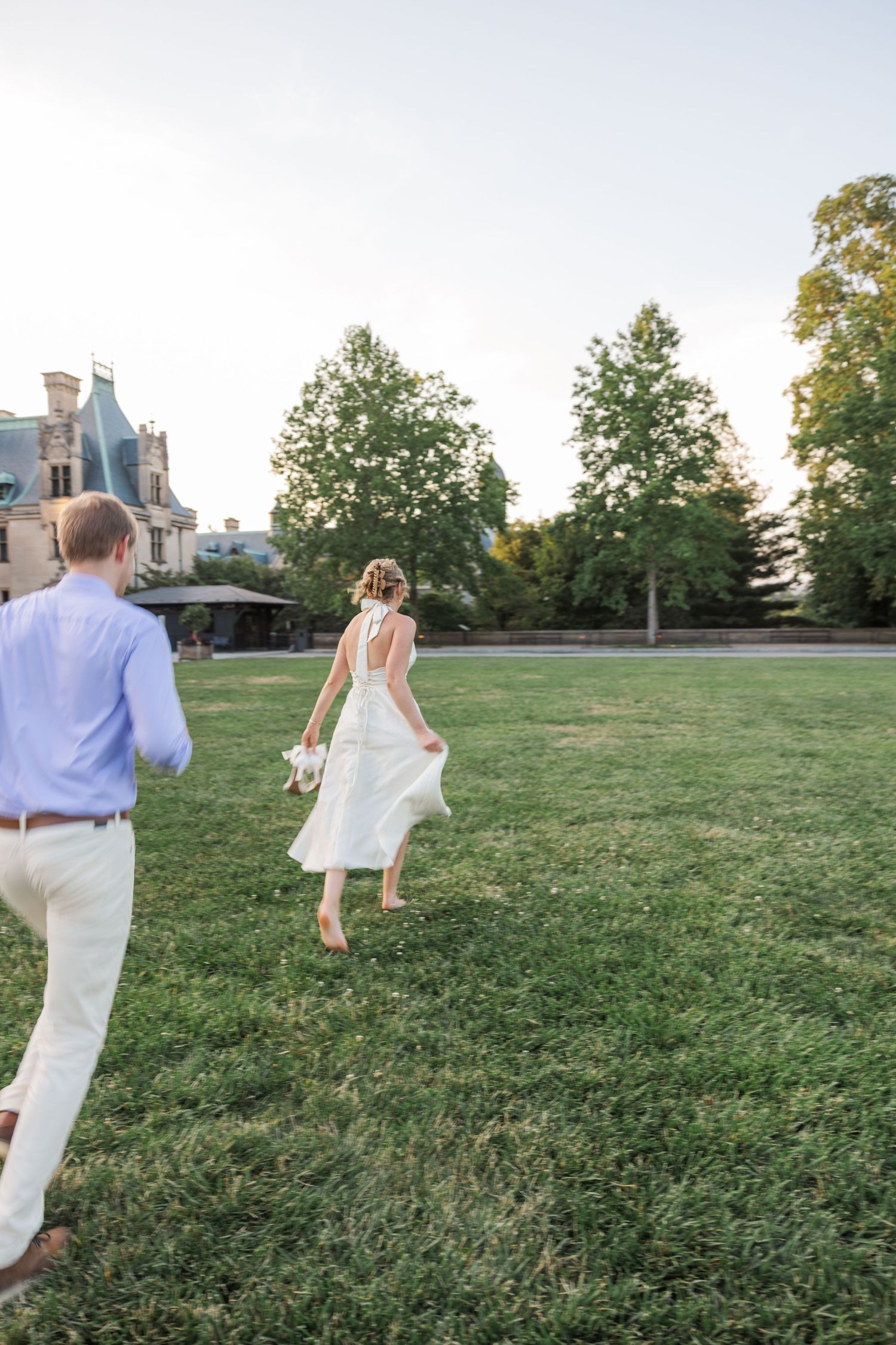 Documentary Wedding Photographer. A couple running playfully across a grassy hilltop with historic buildings visible in the background.