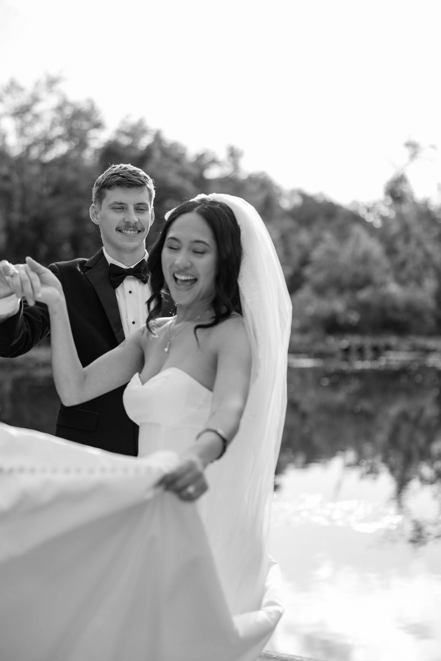 A joyful black and white wedding photo showing newlyweds dancing by a calm lake.