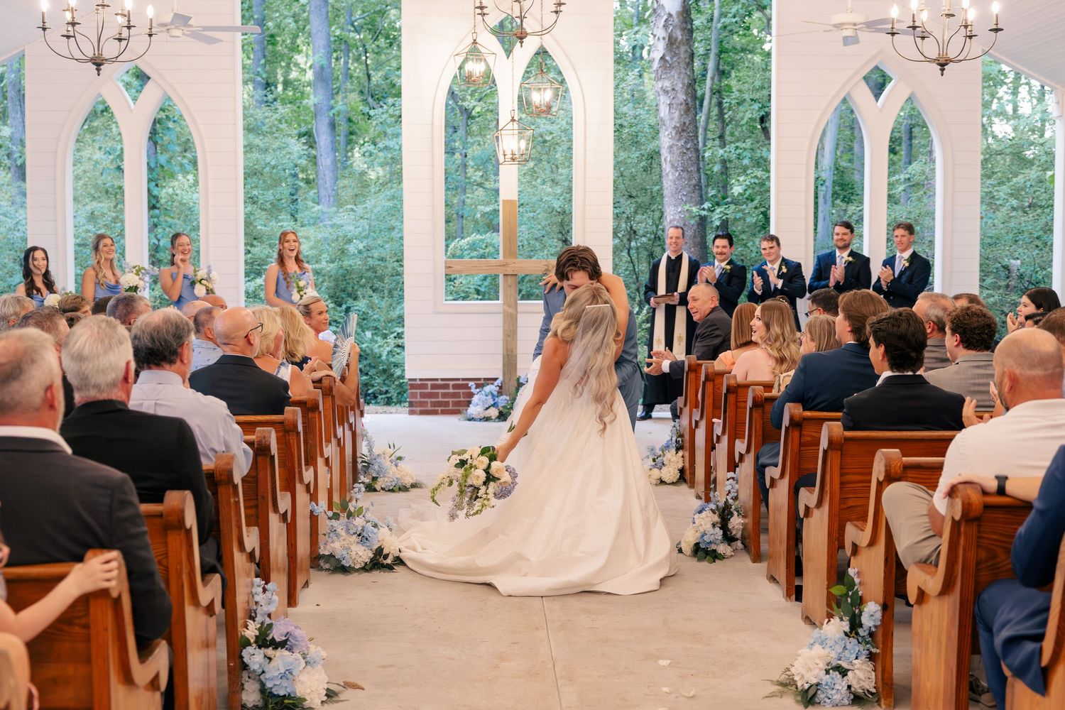 Bride walks down aisle in sunlit chapel with arched windows and wooden pews filled with wedding guests.