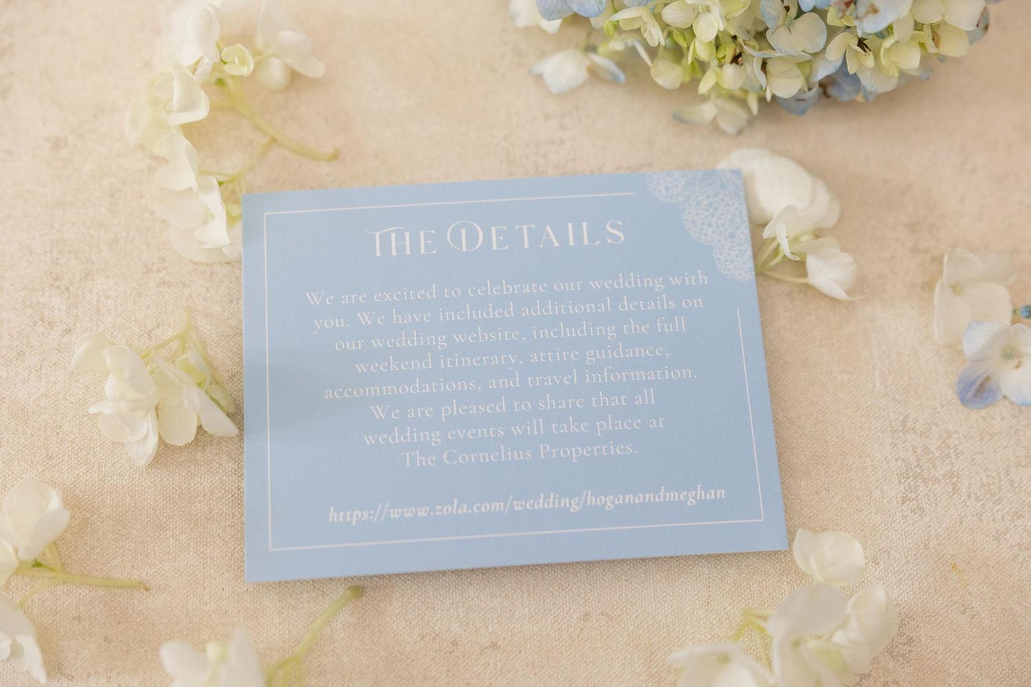 Elegant dusty blue wedding details card surrounded by white hydrangea flowers on a neutral background.