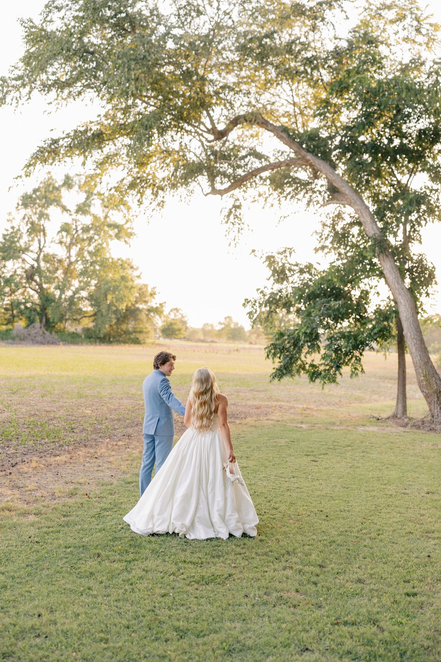 Bride and groom walking hand in hand across a lush green field during golden hour at their outdoor wedding.