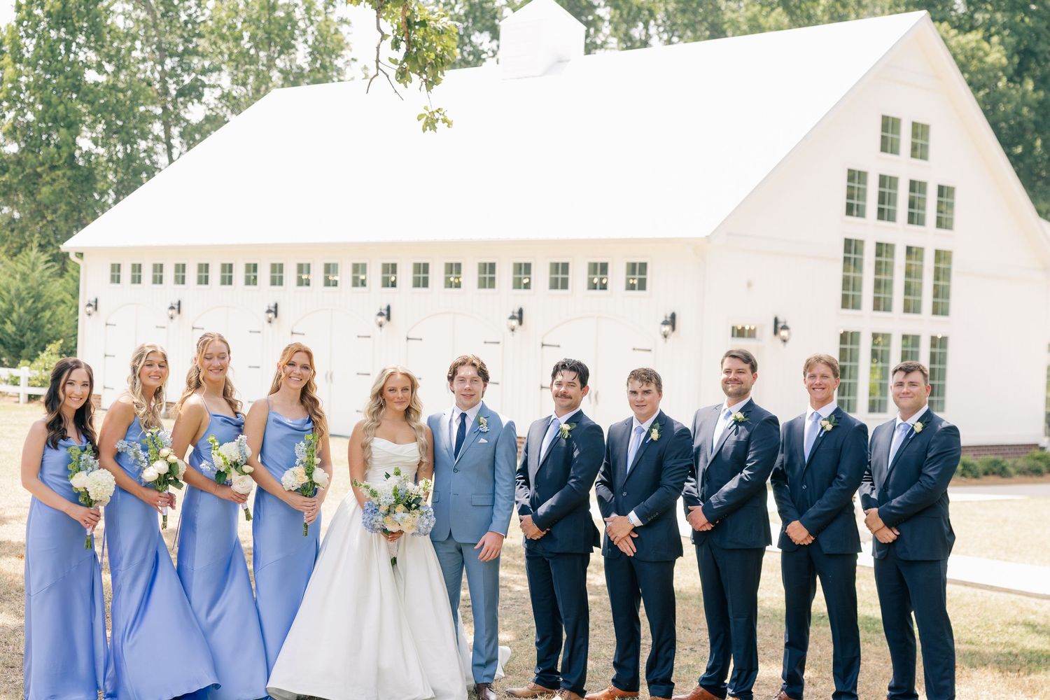 Wedding party posing in front of a white barn with bridesmaids in blue dresses and groomsmen in navy.