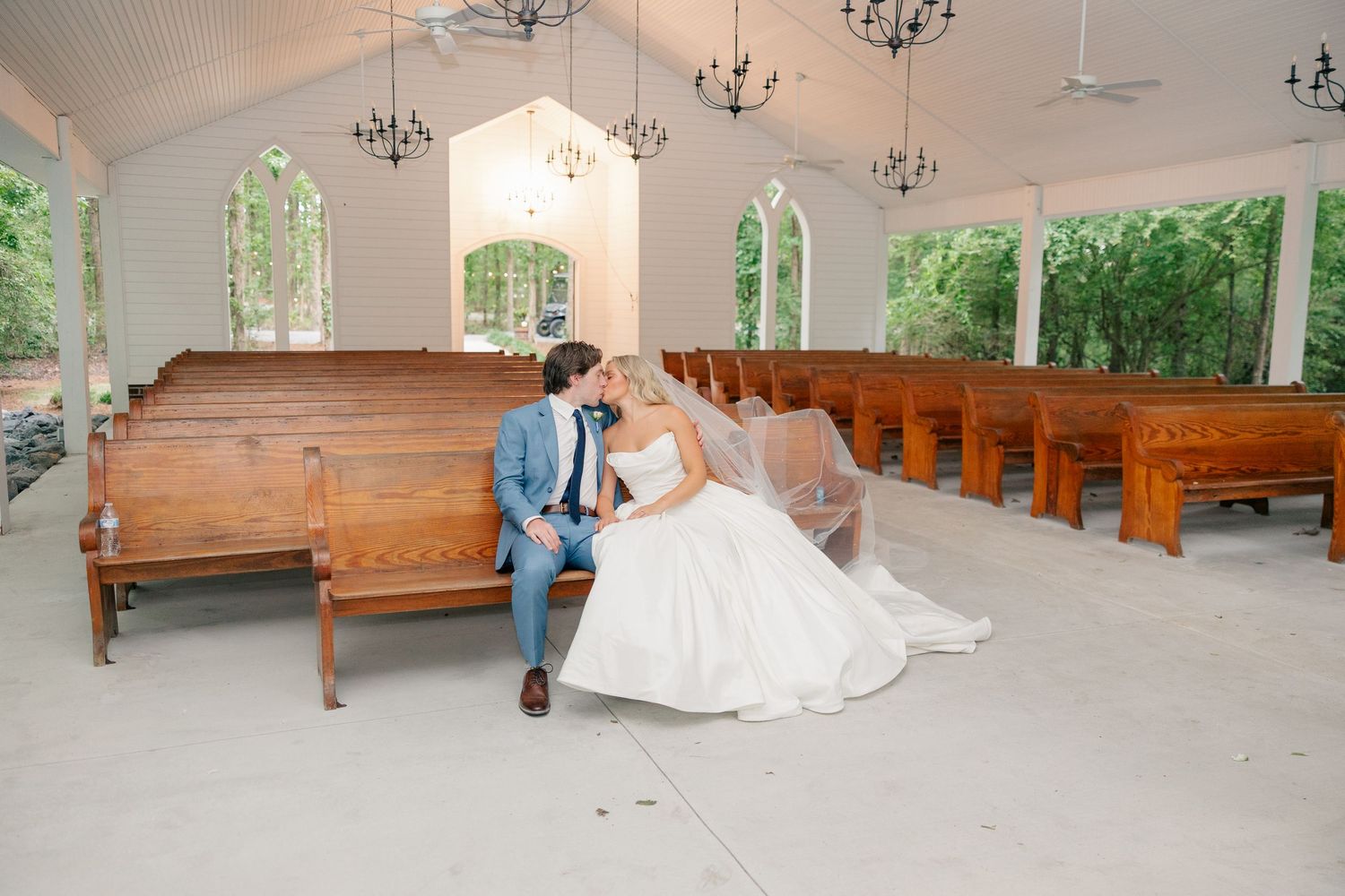 Bride and groom sharing a kiss while seated on a wooden pew in a white chapel.