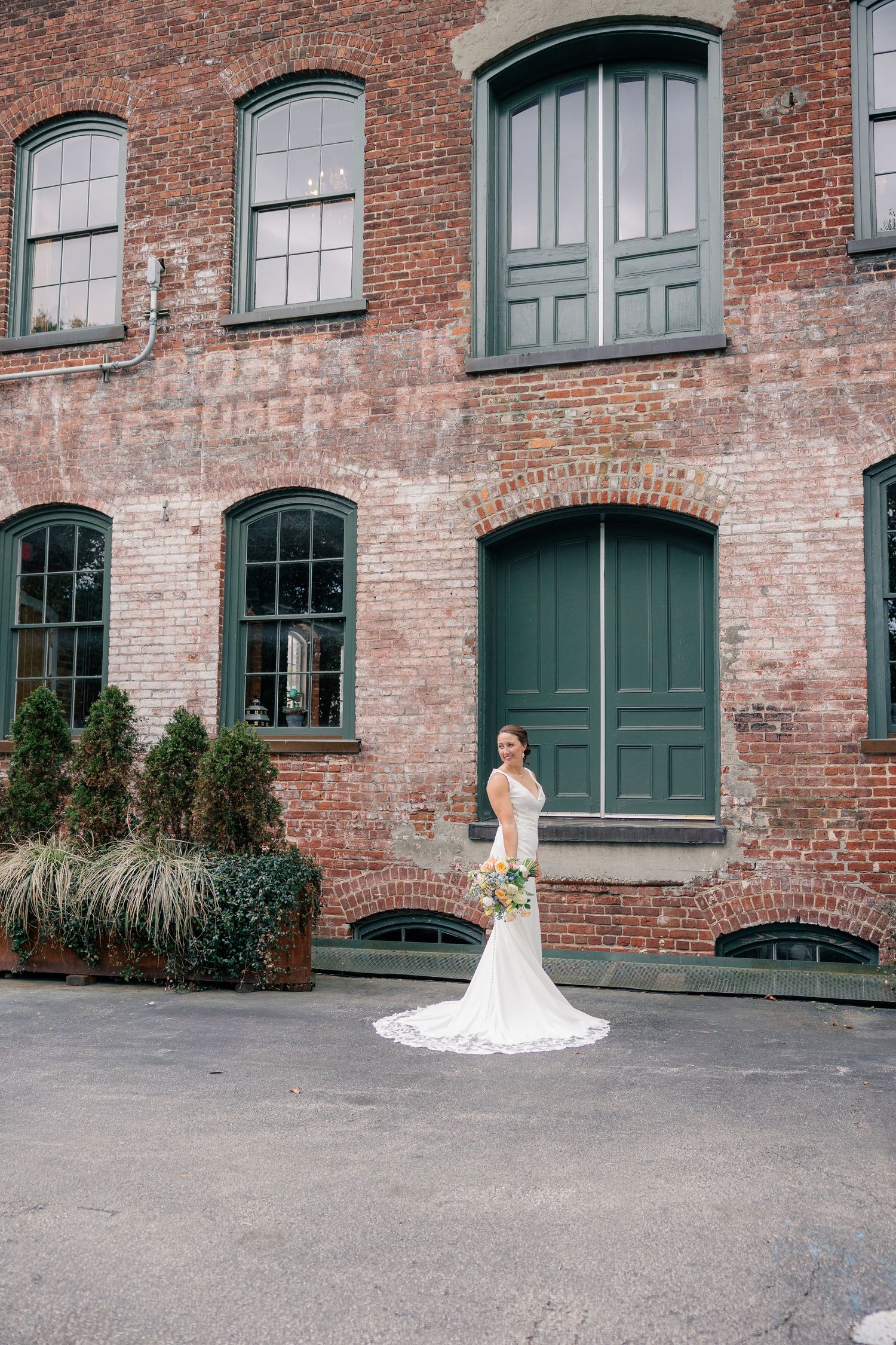Elegant bride in a lace gown poses alone in front of a historic rustic red brick building.