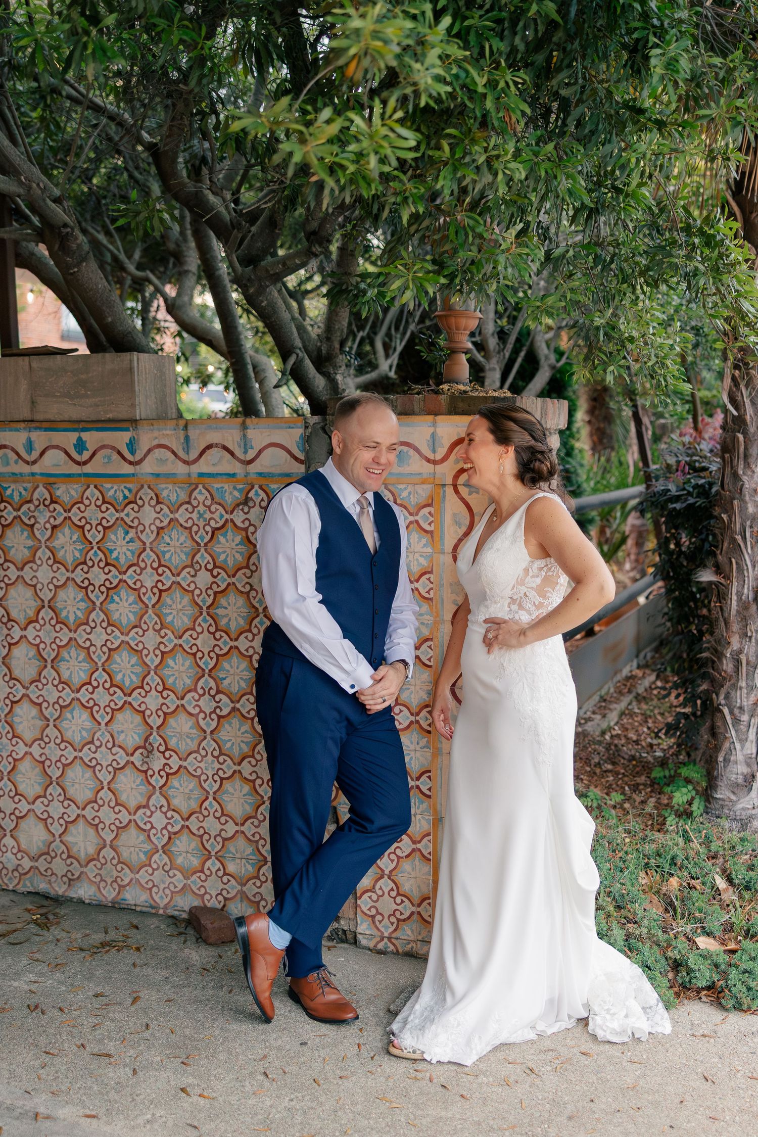 Bride and groom share a joyful moment beside a decorative tiled wall surrounded by lush greenery.