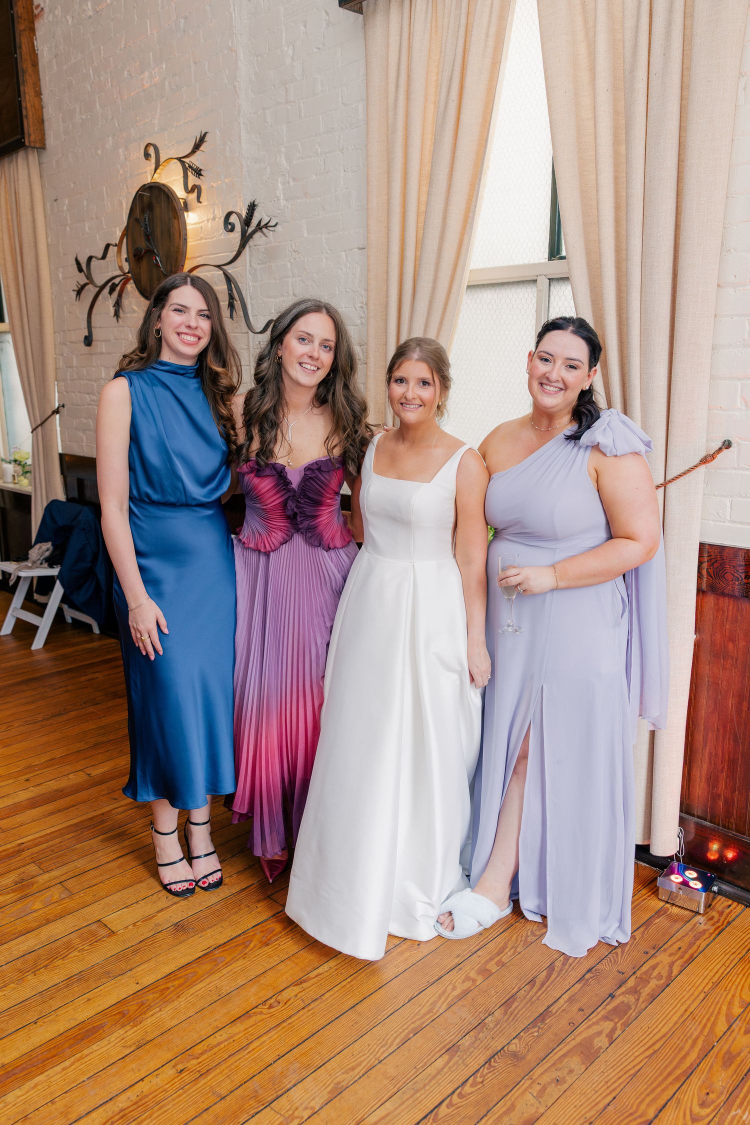 Four women pose together at a wedding, including a bride in white and three bridesmaids in blue, purple, and lavender gowns.