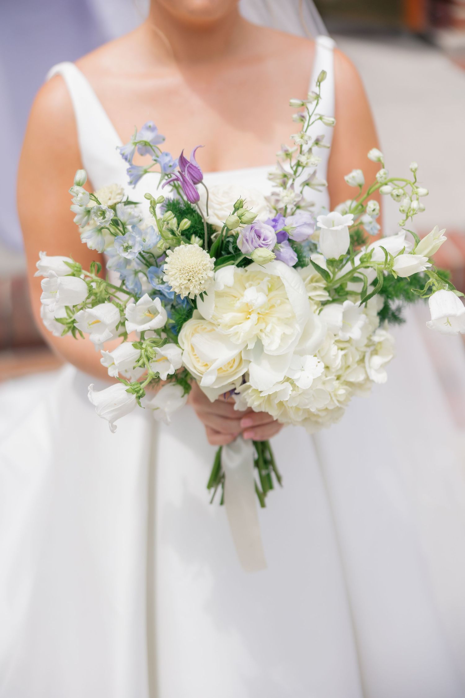Bride holds elegant white and lavender wedding bouquet with peonies and delphinium blooms.