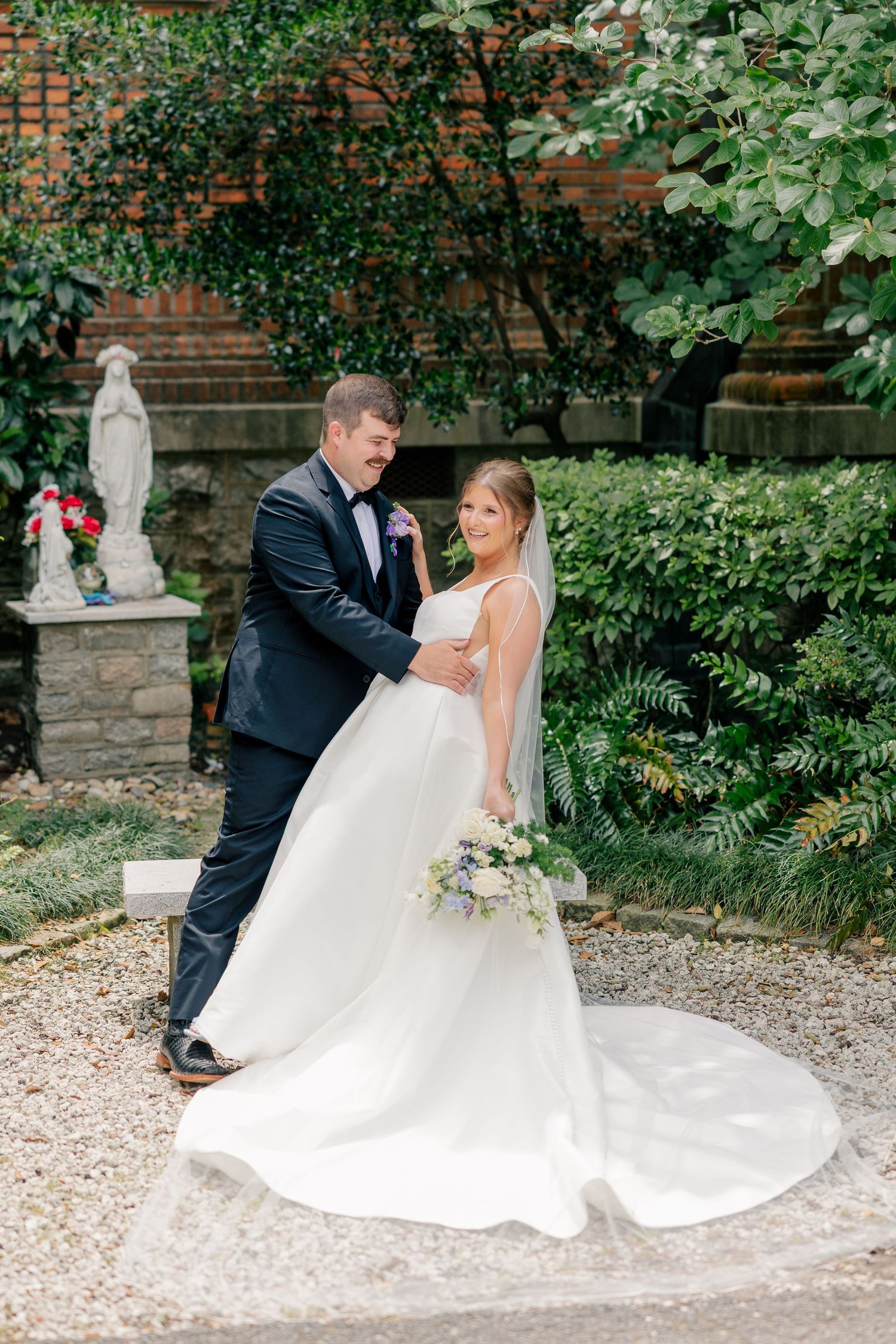 Bride and groom embracing in a lush garden courtyard with a white statue in the background.