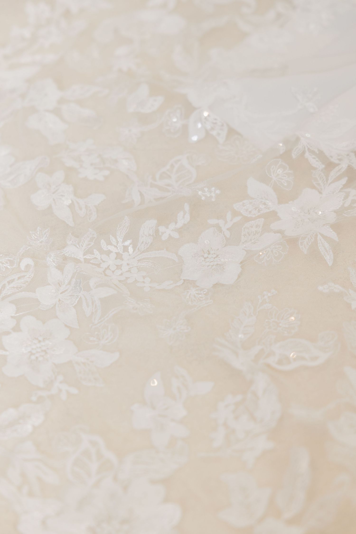 Close-up of delicate white floral lace fabric with intricate embroidery and subtle sparkle details on ivory background.