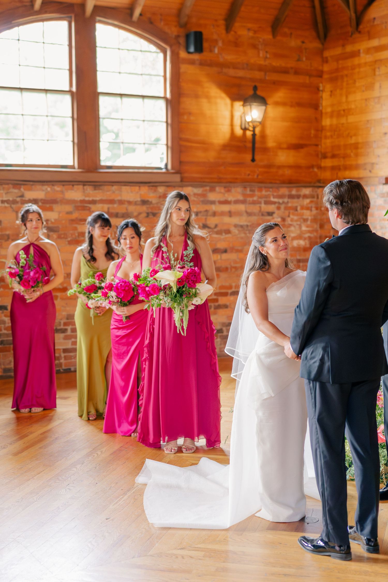 Bride and groom exchanging vows with bridesmaids in fuchsia and green gowns in rustic brick venue.