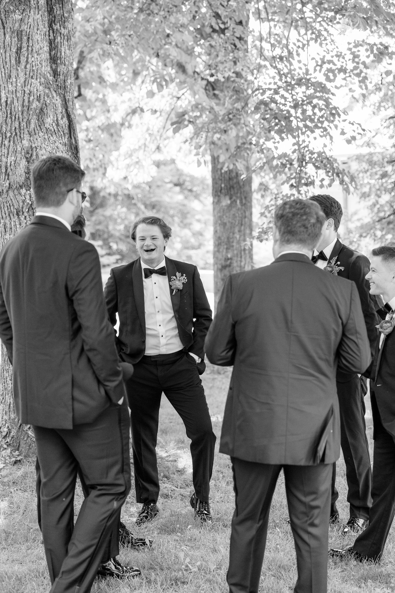 Groomsmen in tuxedos laughing together outdoors under trees in a candid black and white wedding photo.