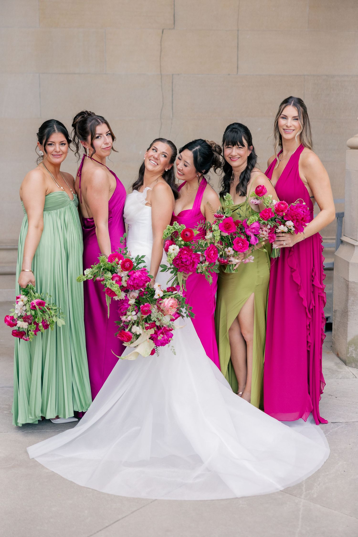 Bride in white gown poses with bridesmaids in pink and green dresses holding vibrant bouquets.