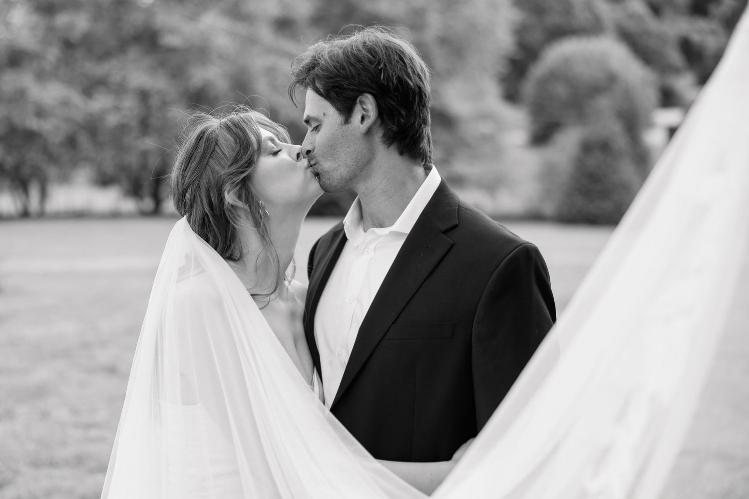 Black and white portrait of bride and groom sharing a romantic kiss beneath flowing veil.