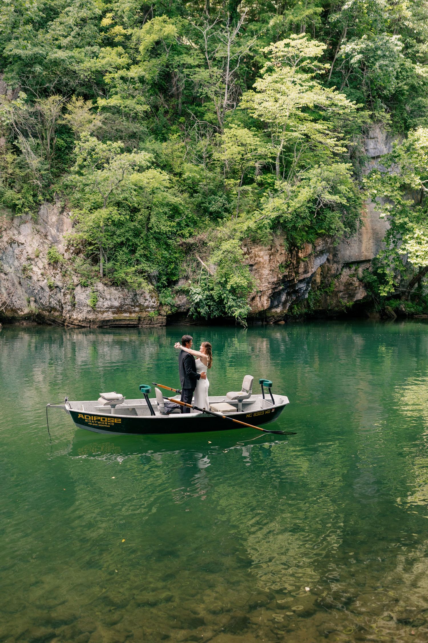 Bride and groom embracing while standing in a rowboat on a lush green quarry lake.