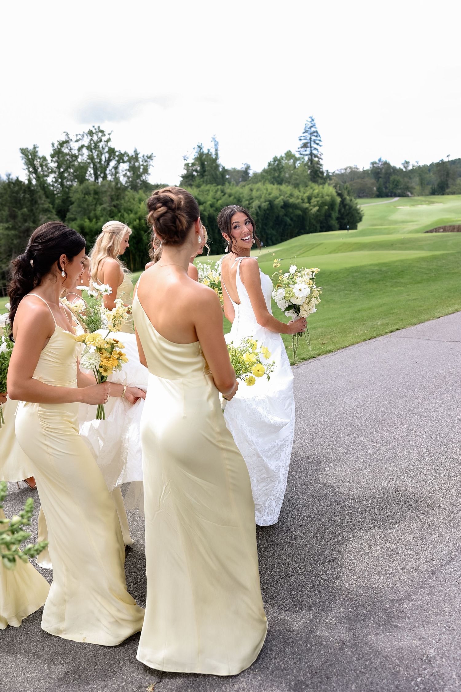 Bridesmaids in ivory gowns holding yellow bouquets walk with a bride in white on a golf course path.