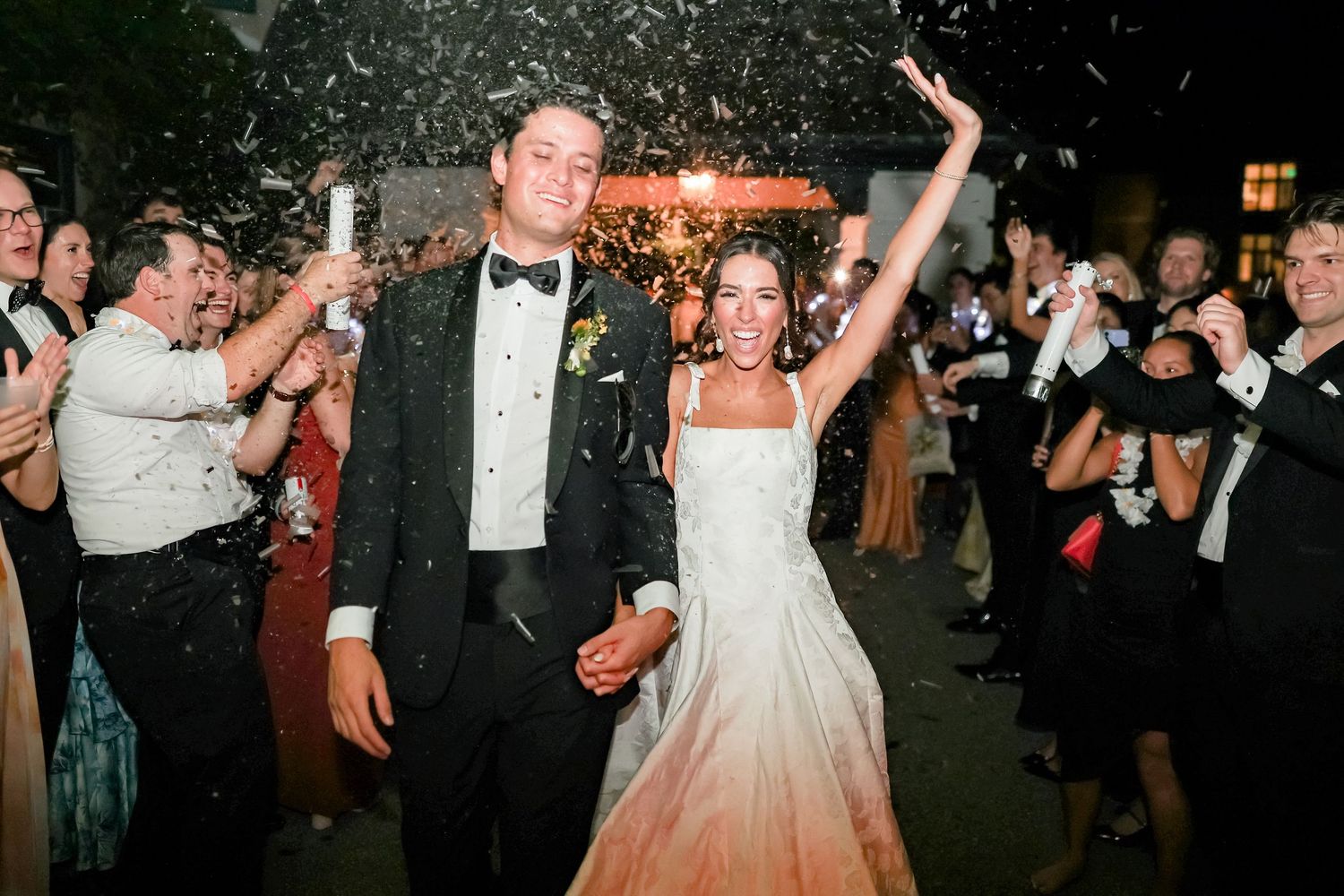 Bride and groom exit wedding reception through a confetti shower, with guests celebrating around them at night.