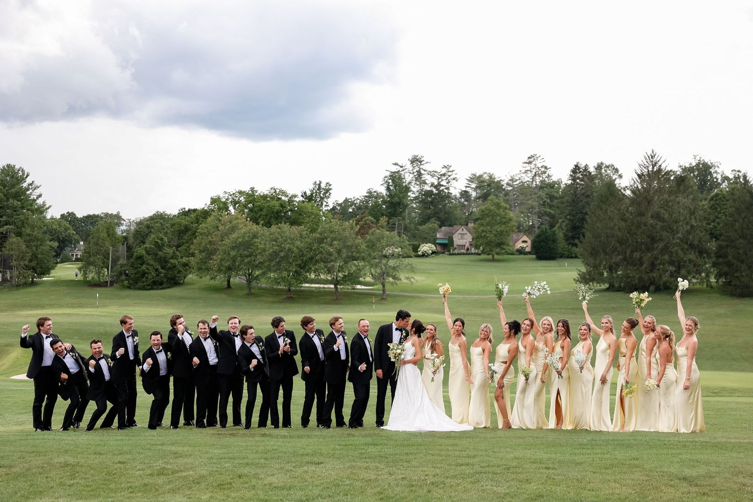 Wedding party on golf course, bride and groom kissing, groomsmen in tuxedos, bridesmaids in gold dresses raising bouquets.