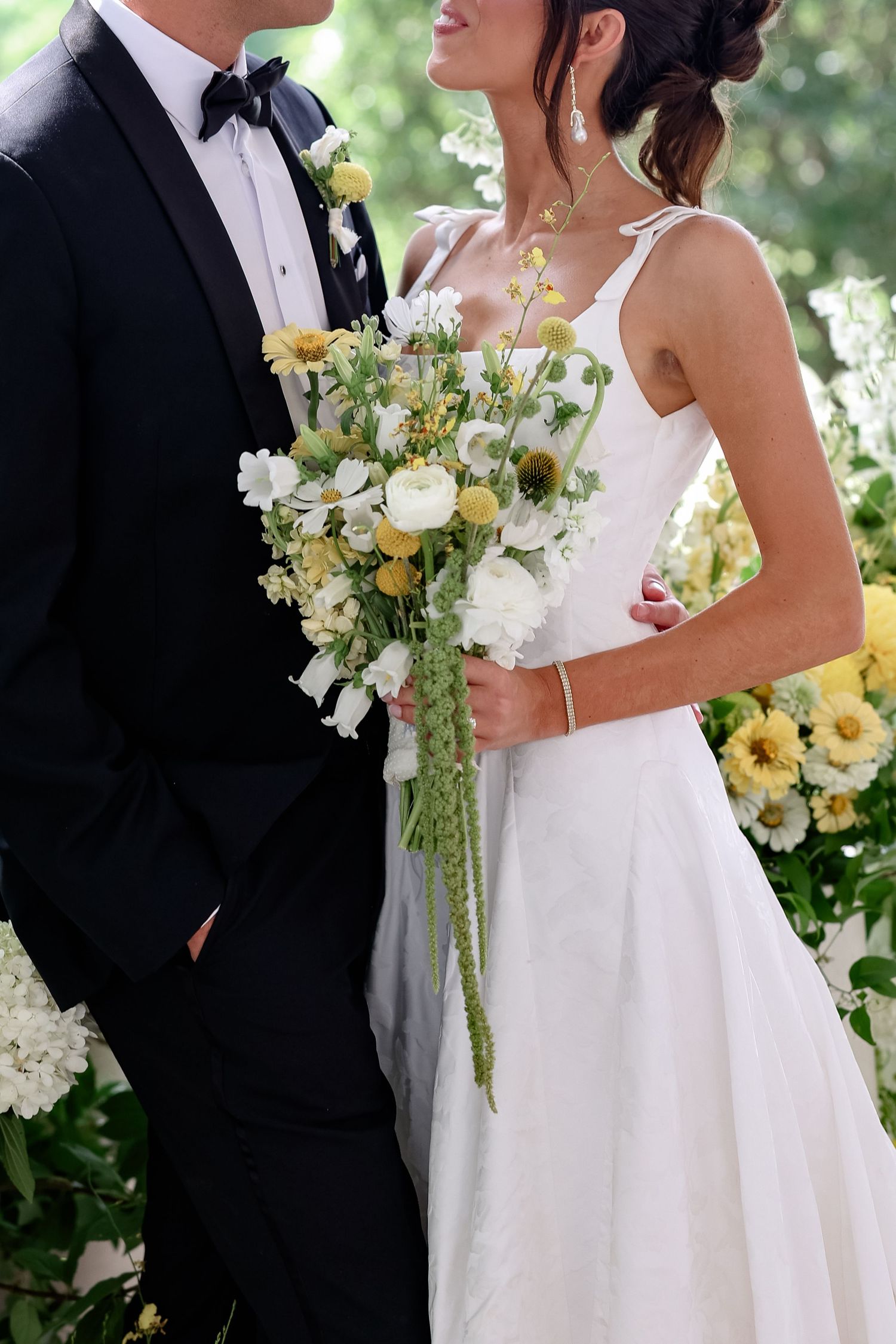 Bride in white gown holds yellow and white floral bouquet while standing with groom in black tuxedo outdoors.