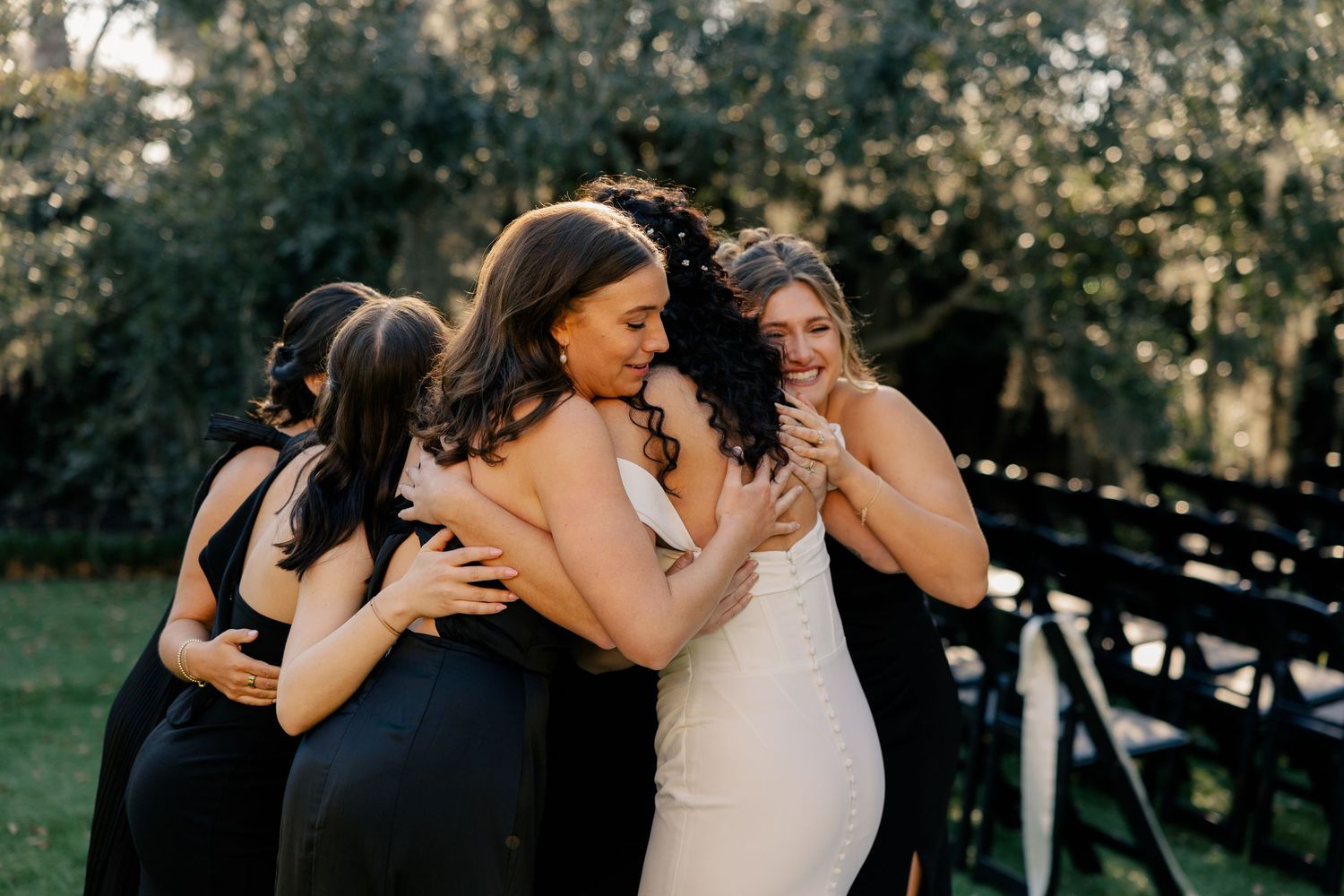 Bridesmaids in black dresses share an emotional group hug with the bride in a white gown outdoors.