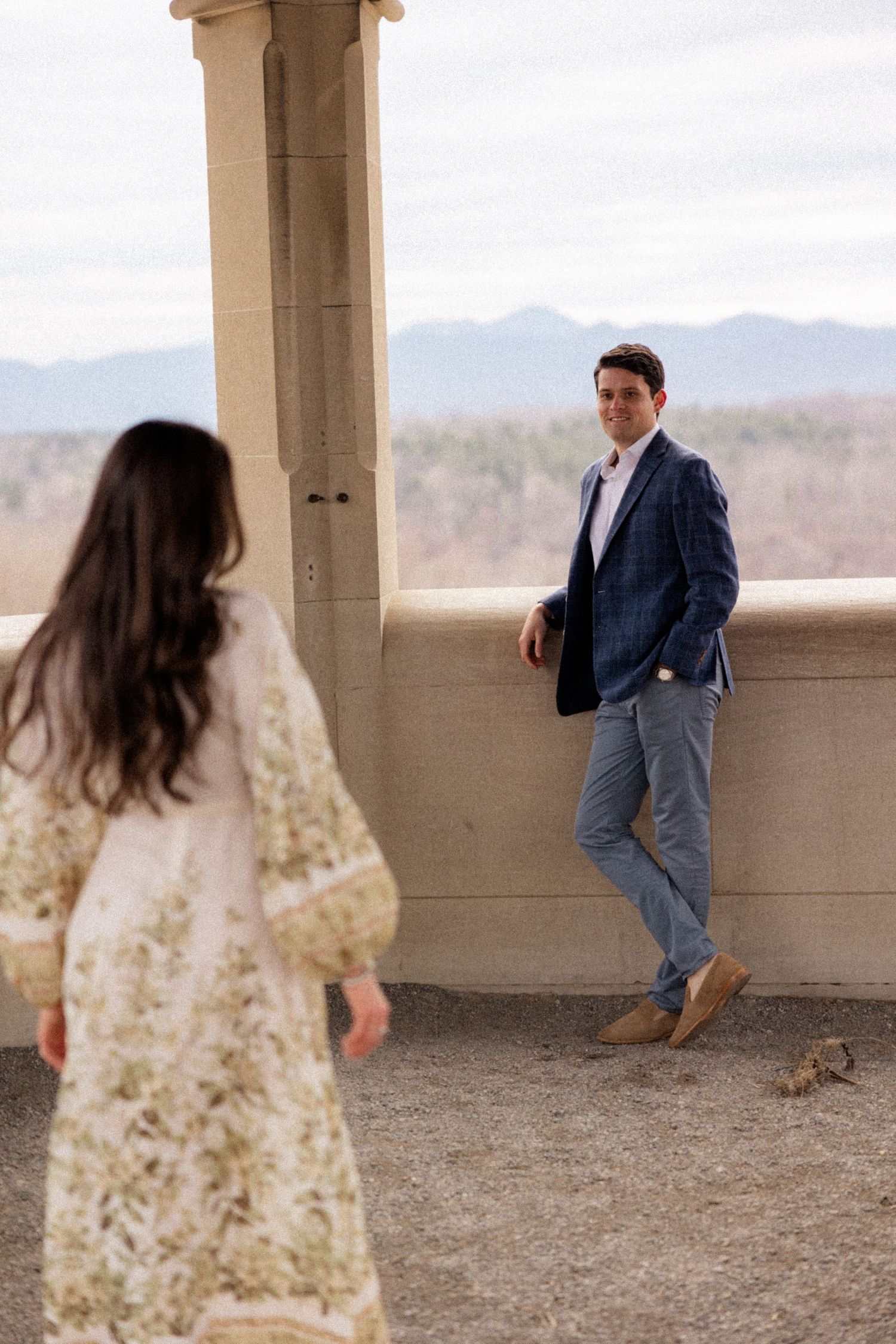 A couple stands on a stone balcony overlooking mountain views, with her in a floral dress and him in a blazer and jeans.