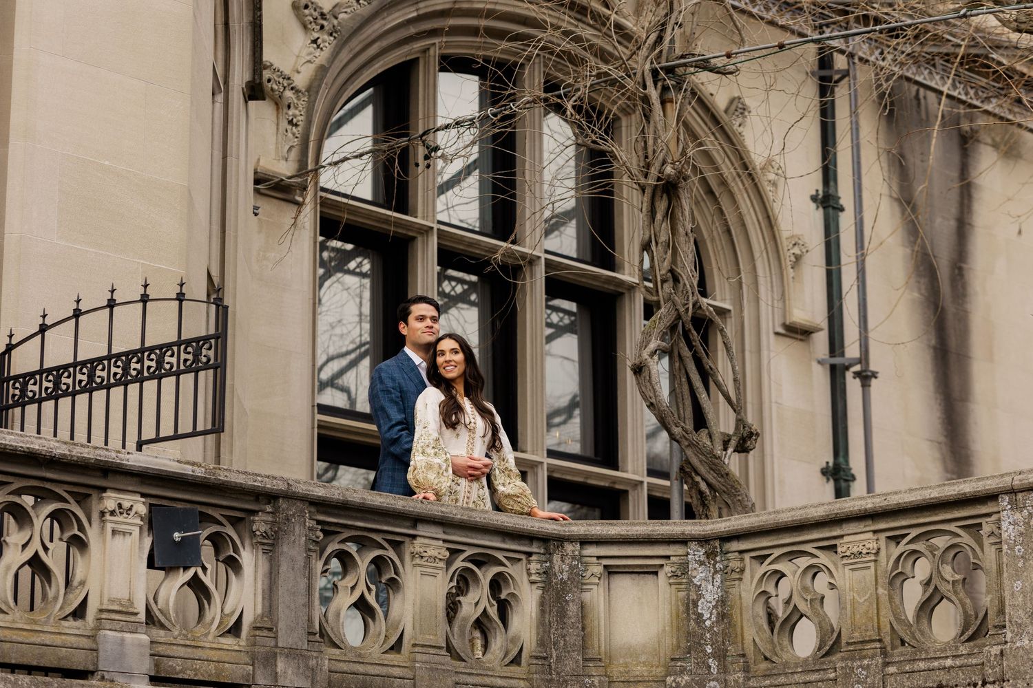 A couple stands on an ornate gothic stone balcony with decorative architectural details and arches.