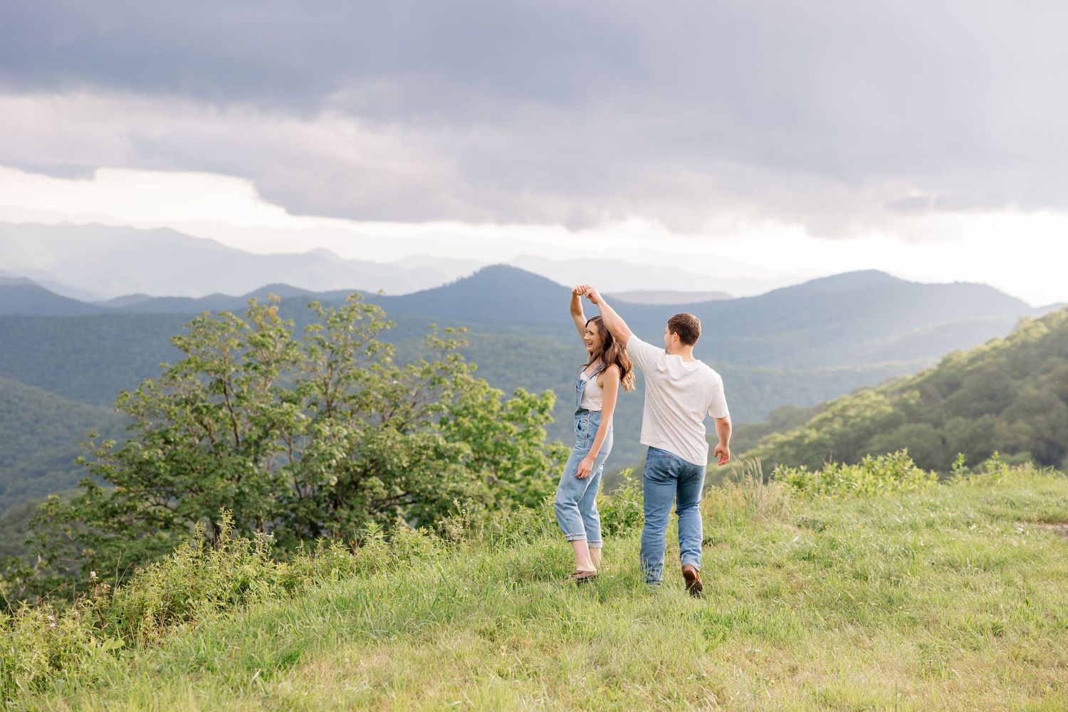 A couple stands together on a scenic mountain overlook with rolling hills and lush greenery stretching into the distance.