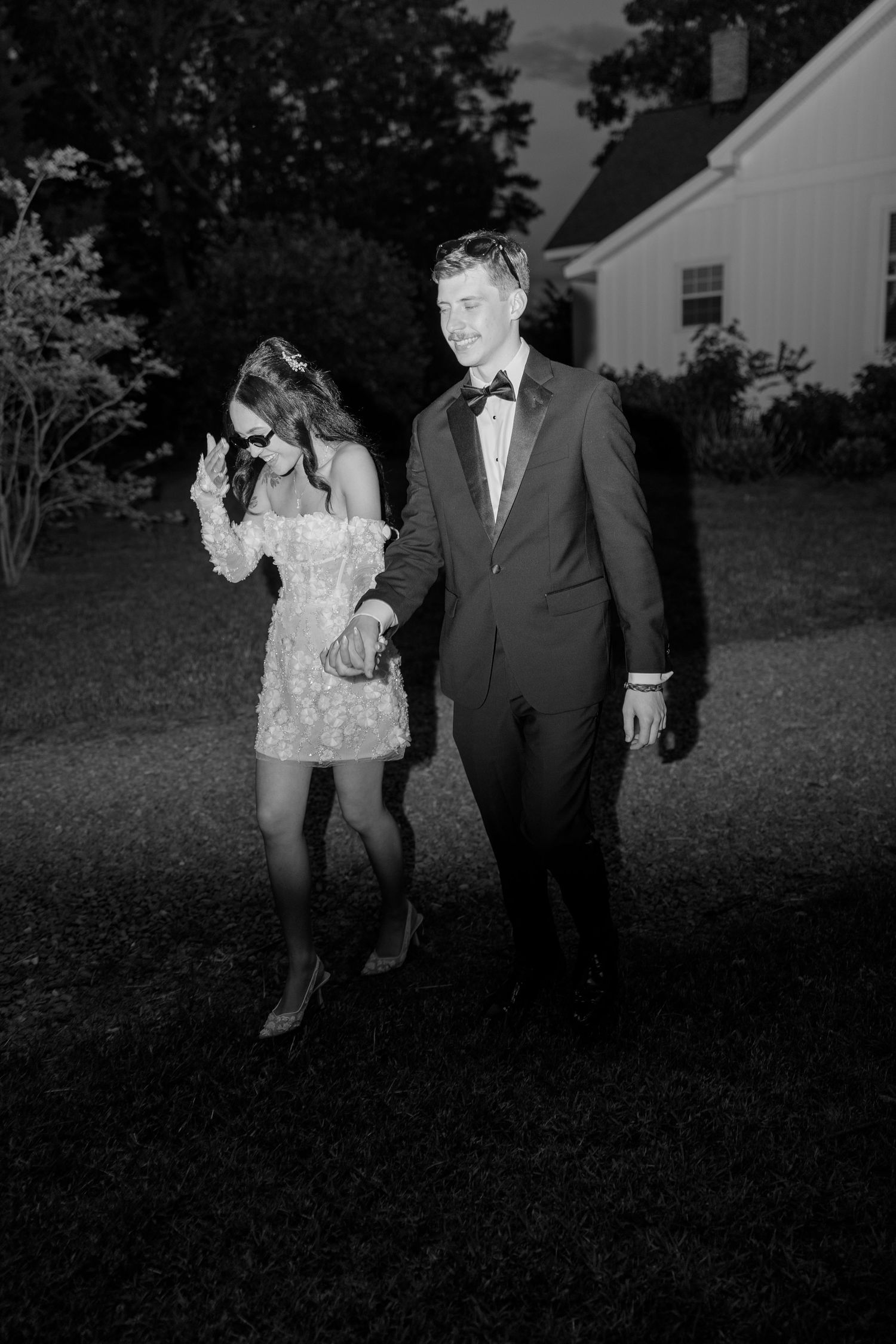 Black and white nighttime portrait showing couple walking across grass outside white building.
