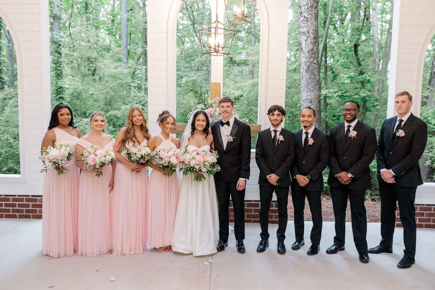Wedding party lined up outside white chapel with bridesmaids in pink and groomsmen in black suits.