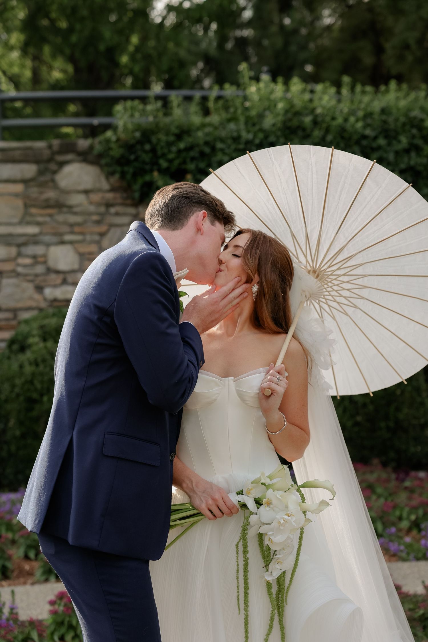 Wedding couple sharing a kiss under a white parasol in a garden.