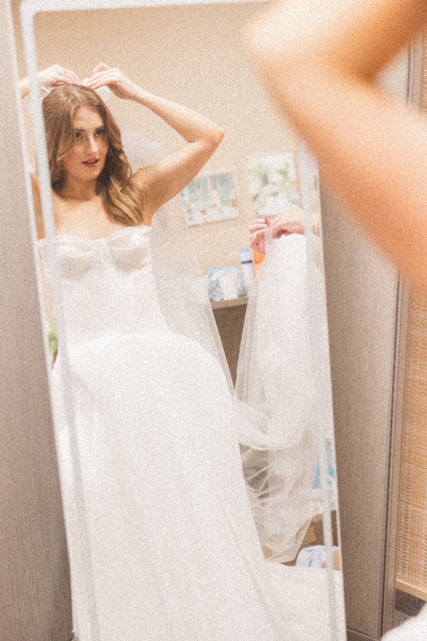 A bride in a white lace wedding dress adjusts her hair while looking in a mirror.