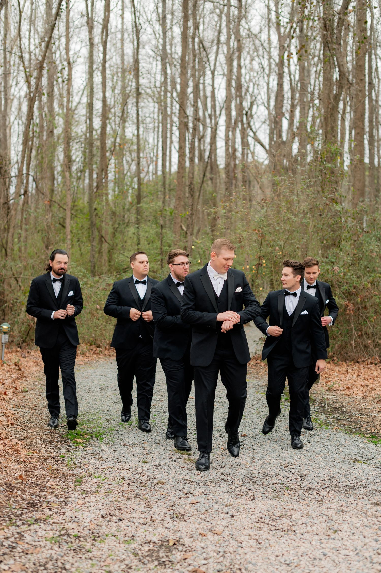 Groomsmen in black tuxedos run playfully down a wooded path covered in fallen leaves.