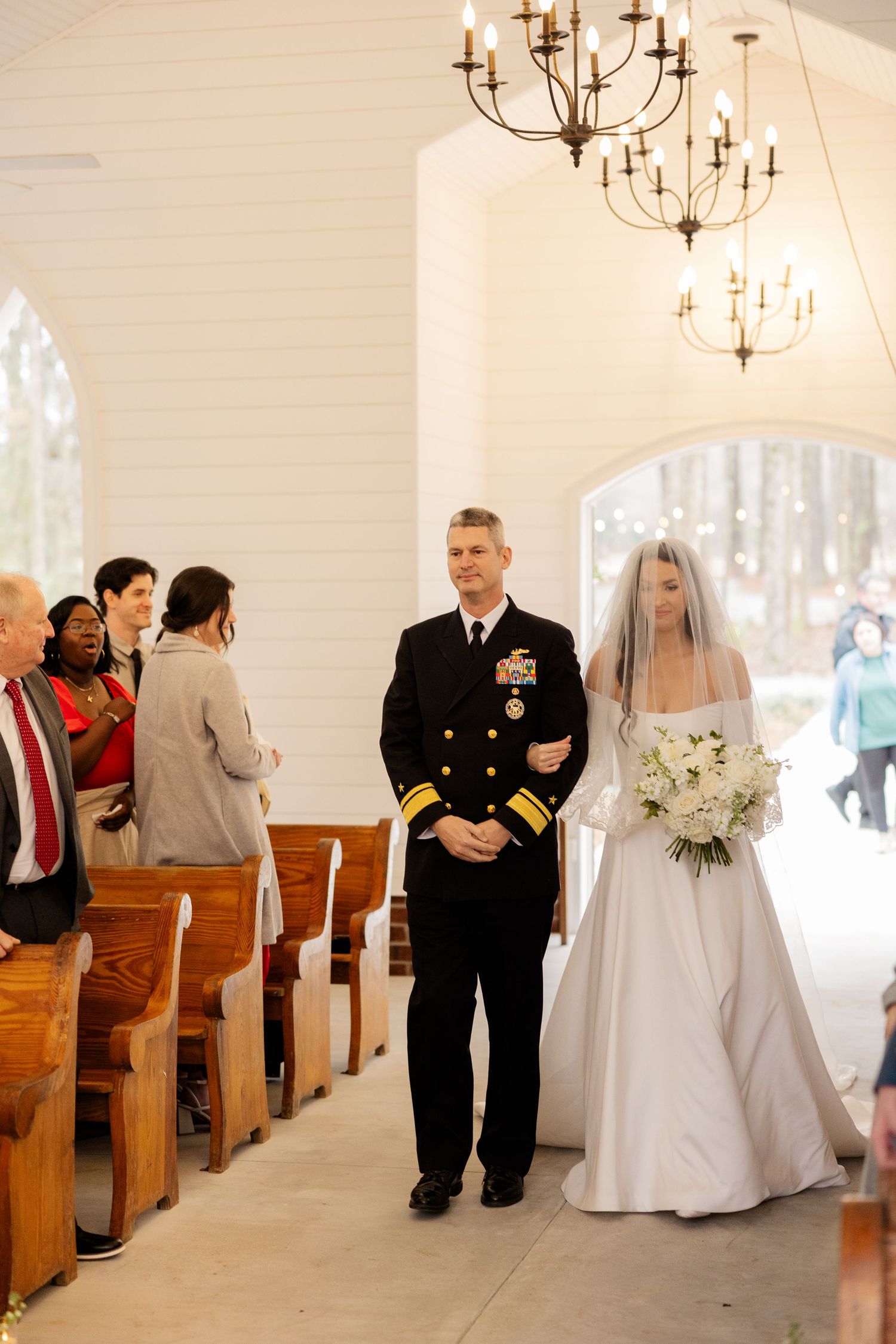 A white chapel interior with wooden pews decorated for a wedding ceremony featuring chandeliers and natural light.