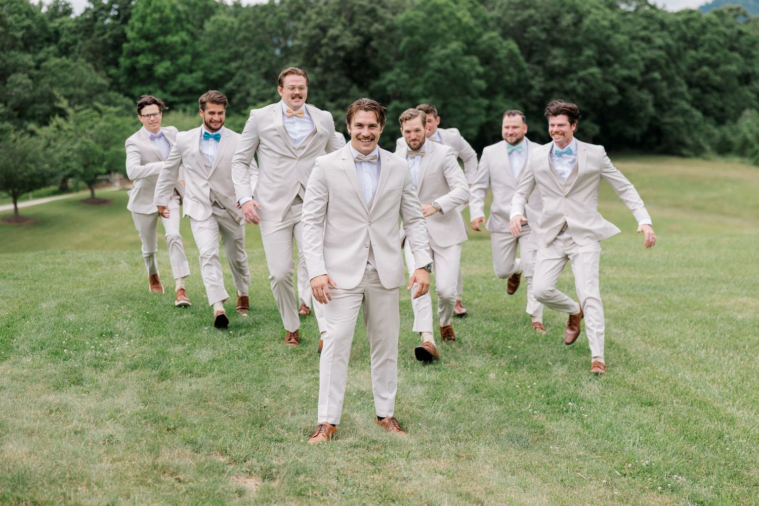 Group in light beige suits running and jumping playfully on green grass.