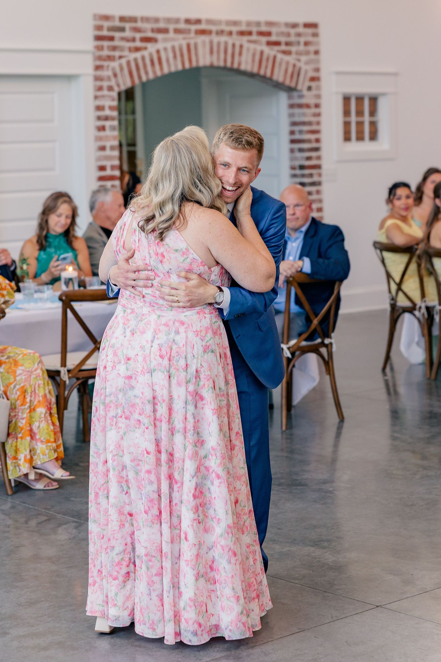 Groom in blue suit shares mother-son dance at elegant indoor wedding reception.