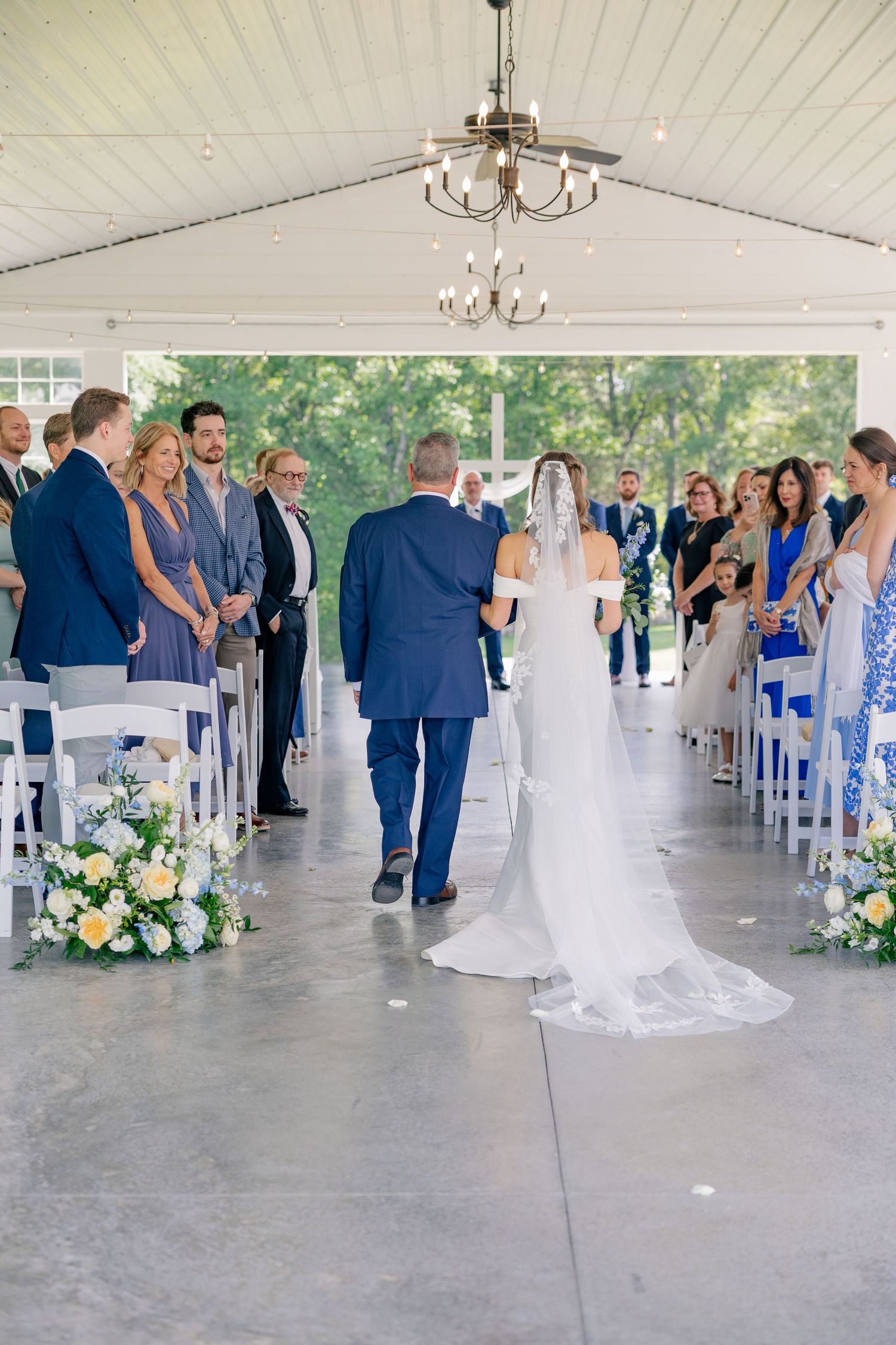 Bride walks down aisle with escort at elegant tented wedding ceremony with floral arrangements.