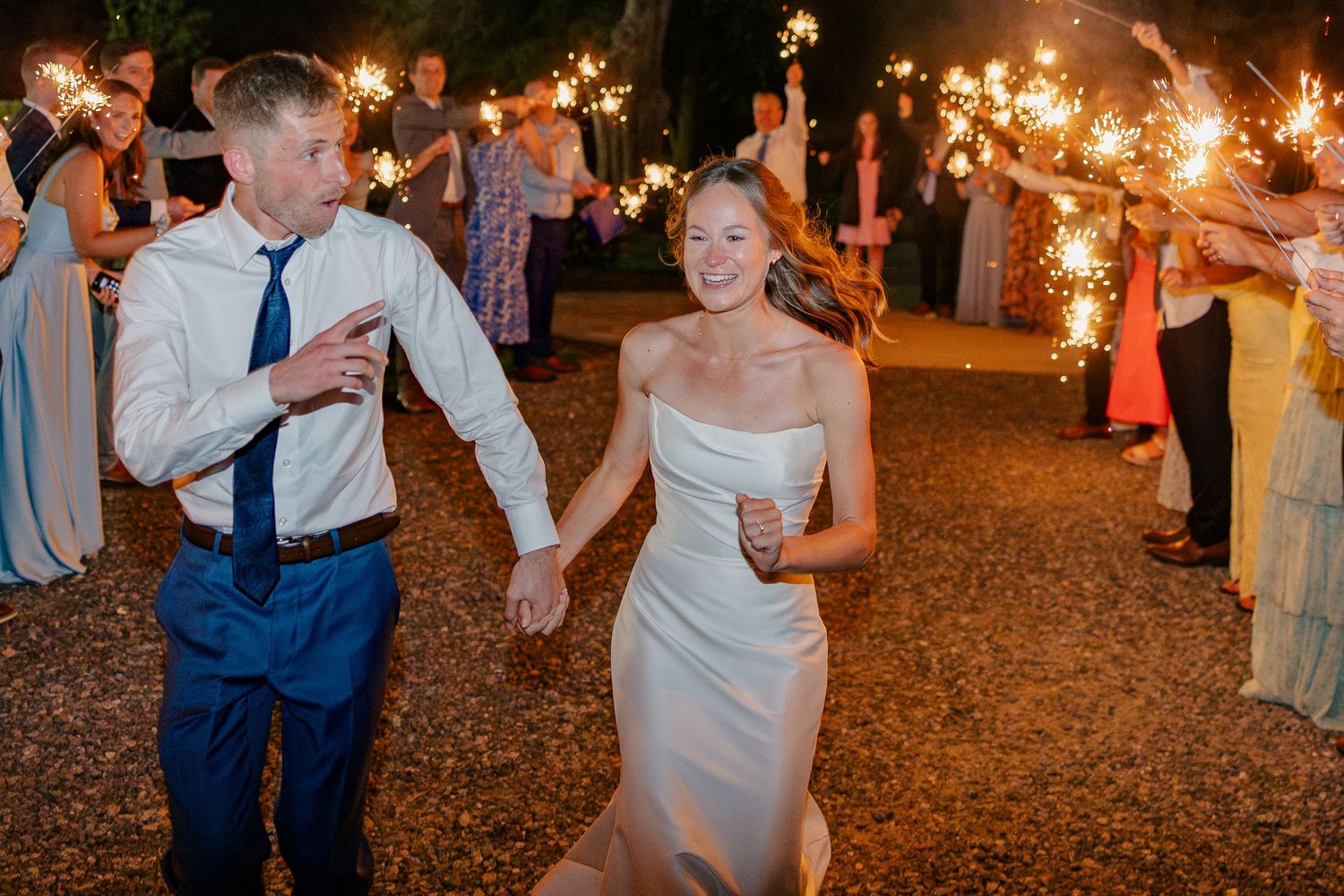 A joyful bride and groom walk hand-in-hand through a sparkler send-off line at their evening wedding.